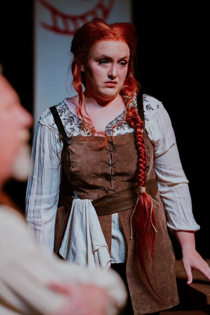 Woman in historical costume with red hair braid, standing in stage setting