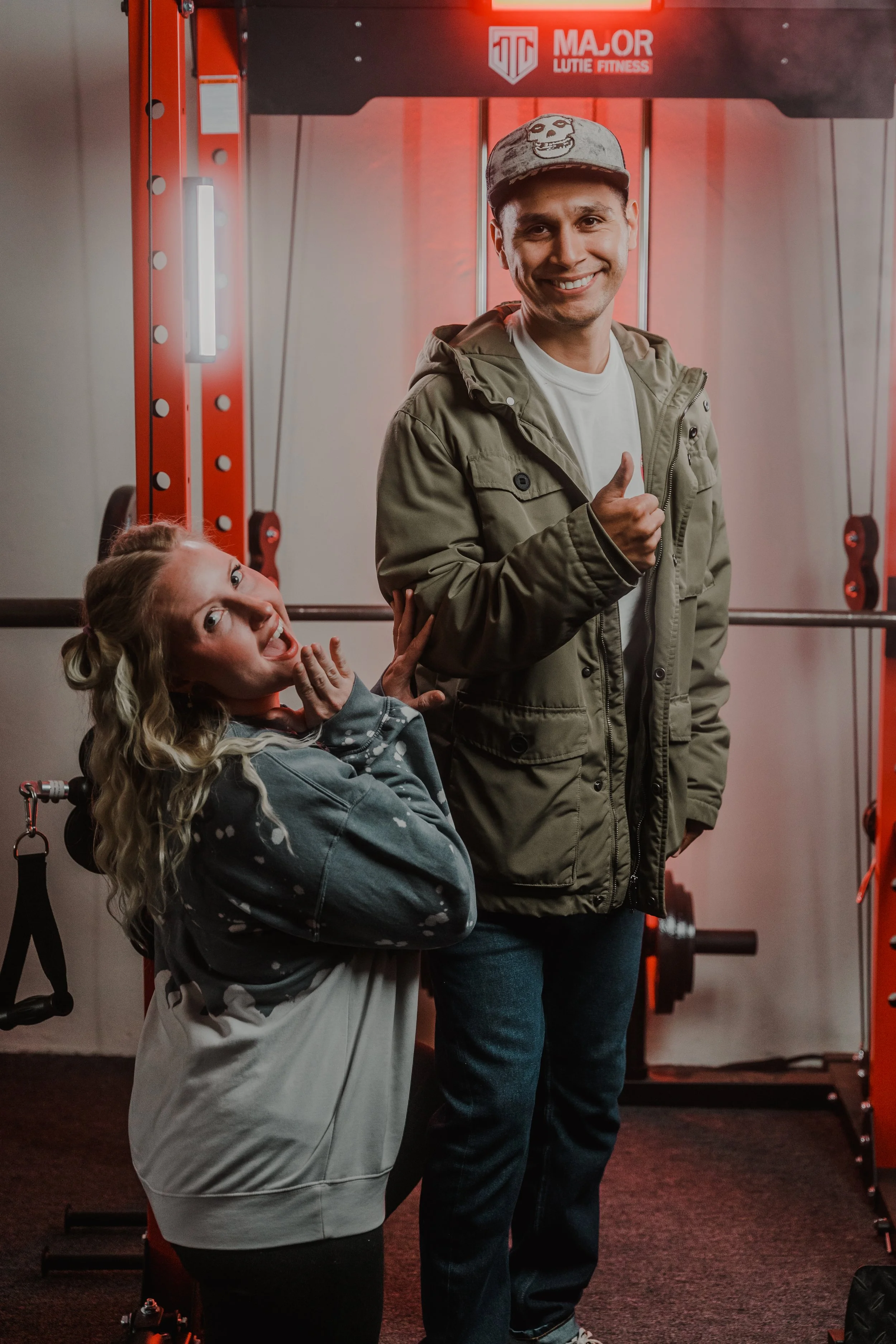 Two people posing in a gym, with fitness equipment in the background. One person is kneeling and smiling, while the other is standing, giving a thumbs-up.