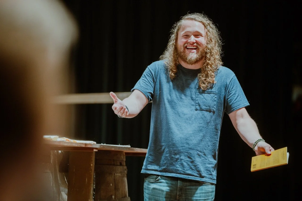 Man with long hair and beard smiling while holding a yellow book on stage.