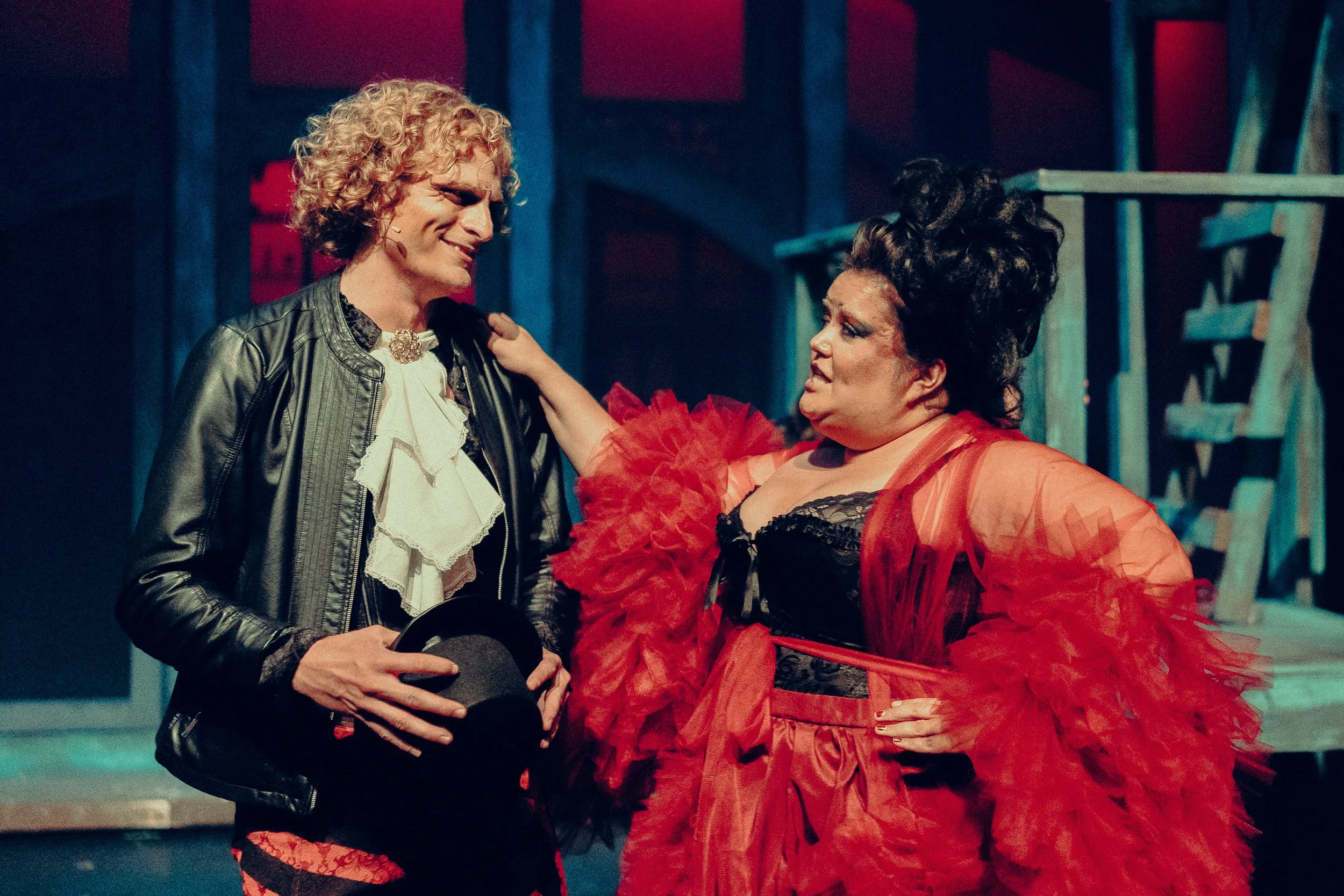 Two performers in theatrical costumes on stage with a dramatic background lighting. One wears a black leather jacket and ruffled shirt, holding a hat, and the other wears a red, feathery outfit.