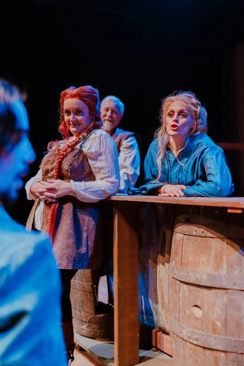 Actors performing on stage in historical costumes, with wooden barrels in the foreground.