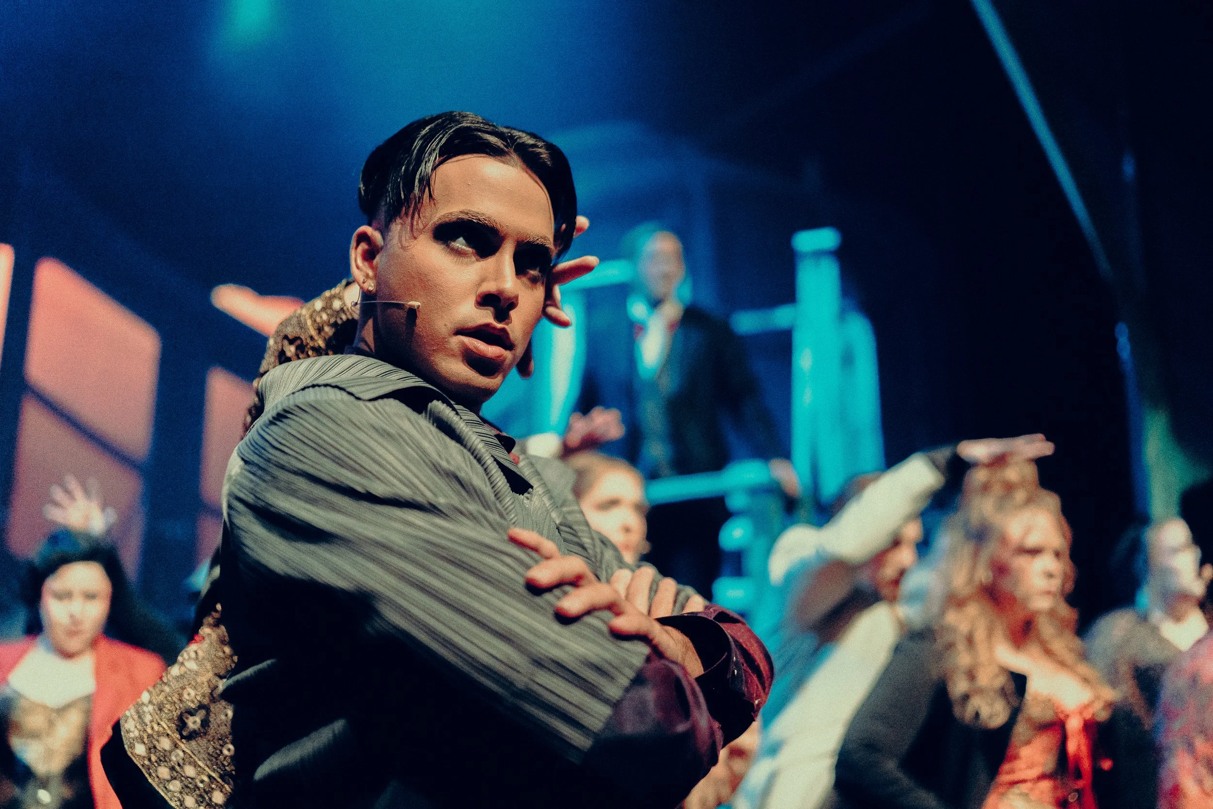 Stage performance with actor in striped suit, surrounded by ensemble cast in colorful costumes, under dramatic lighting.