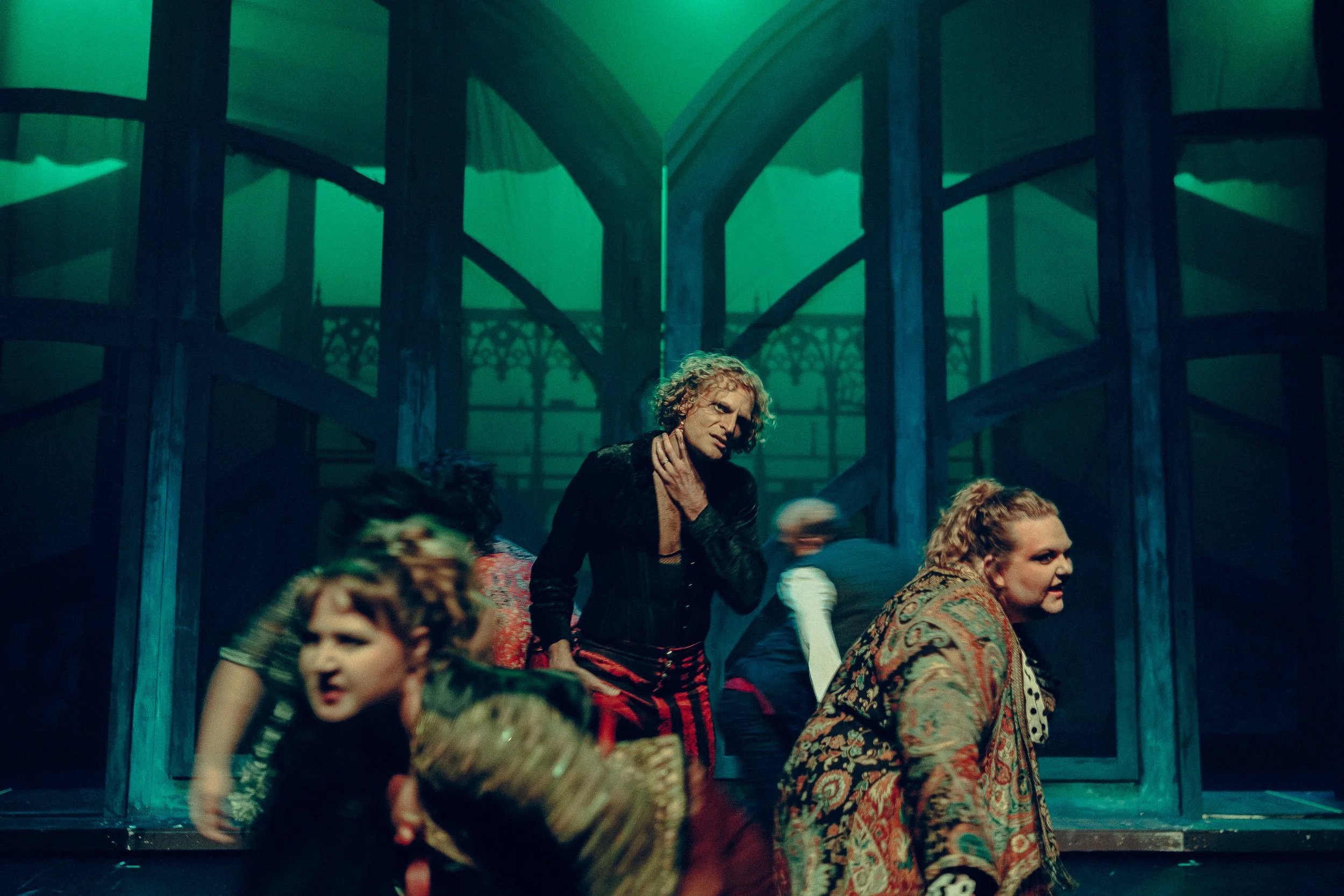 Stage performance with actors in elaborate costumes, set against a dramatic backdrop with an arched design and green lighting.