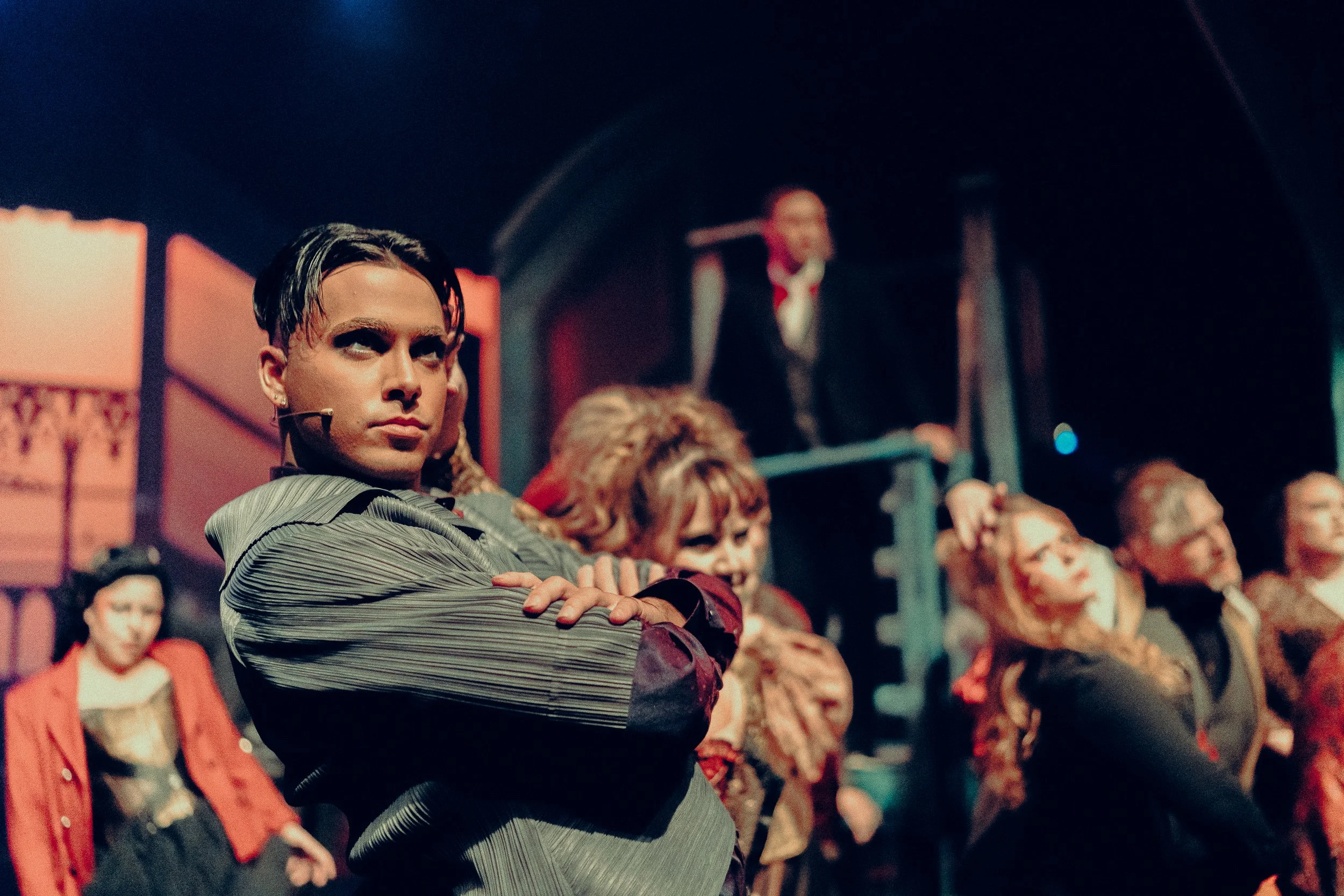 A theatrical performance with multiple actors on stage, one prominently in the foreground looking dramatically towards the side with others blurred in the background. The lighting is warm and dramatic.