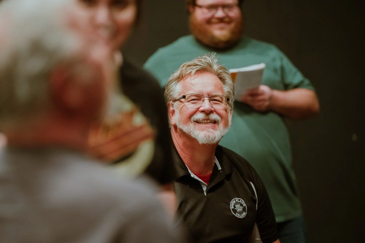 Group of people smiling and interacting indoors, focus on a man with glasses and white beard in the center, blurred individuals in the background.