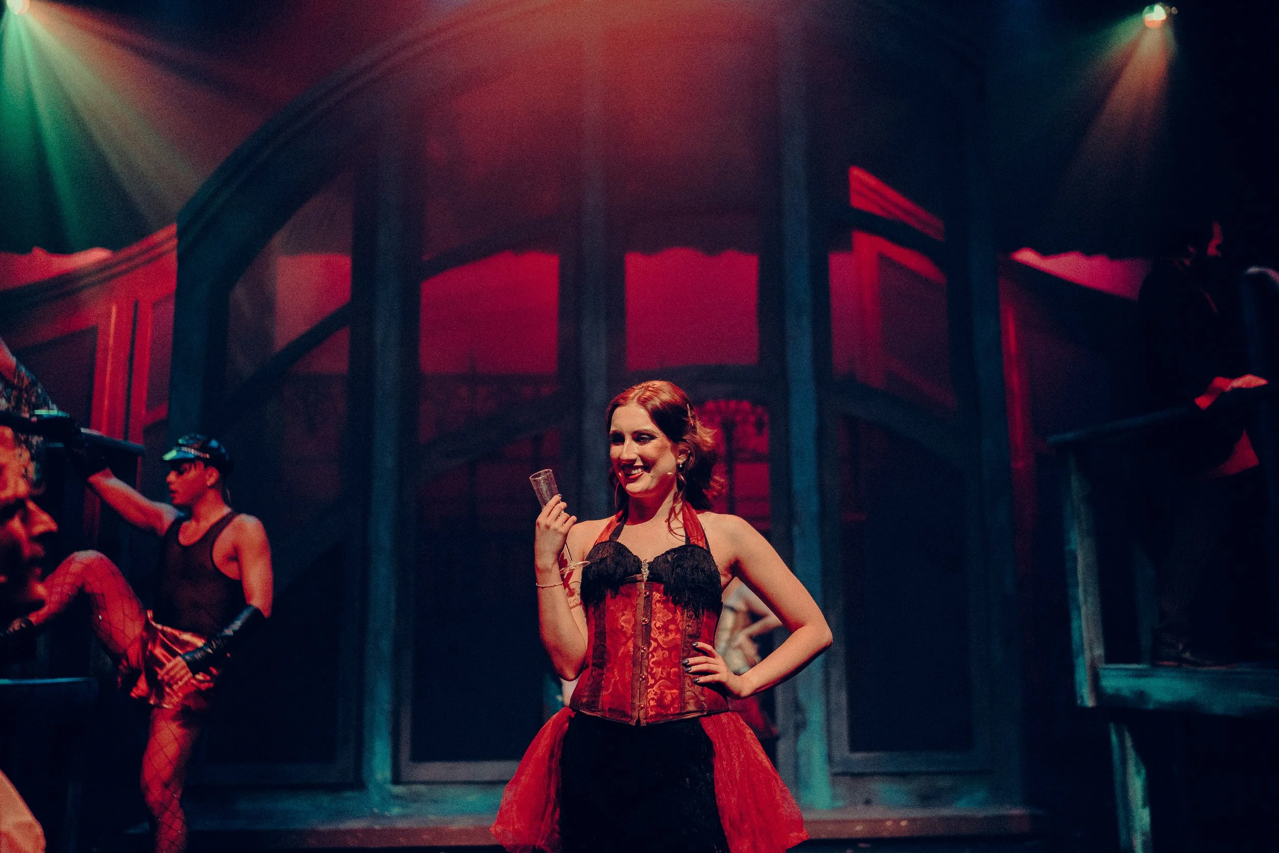 Theatrical performance with a woman in a red and black corset holding a glass on stage, surrounded by performers in a dimly lit setting.