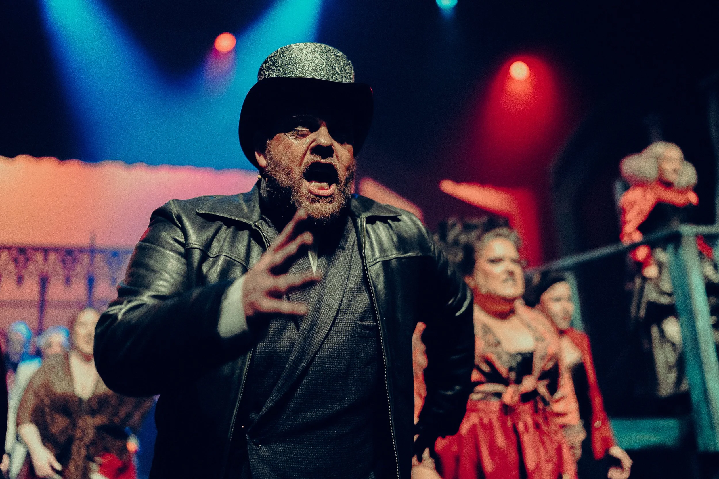 Man performing in a theatrical stage play, wearing a black leather jacket and decorative top hat, with dramatic facial expression, surrounded by other performers in red costumes, under colorful stage lights.