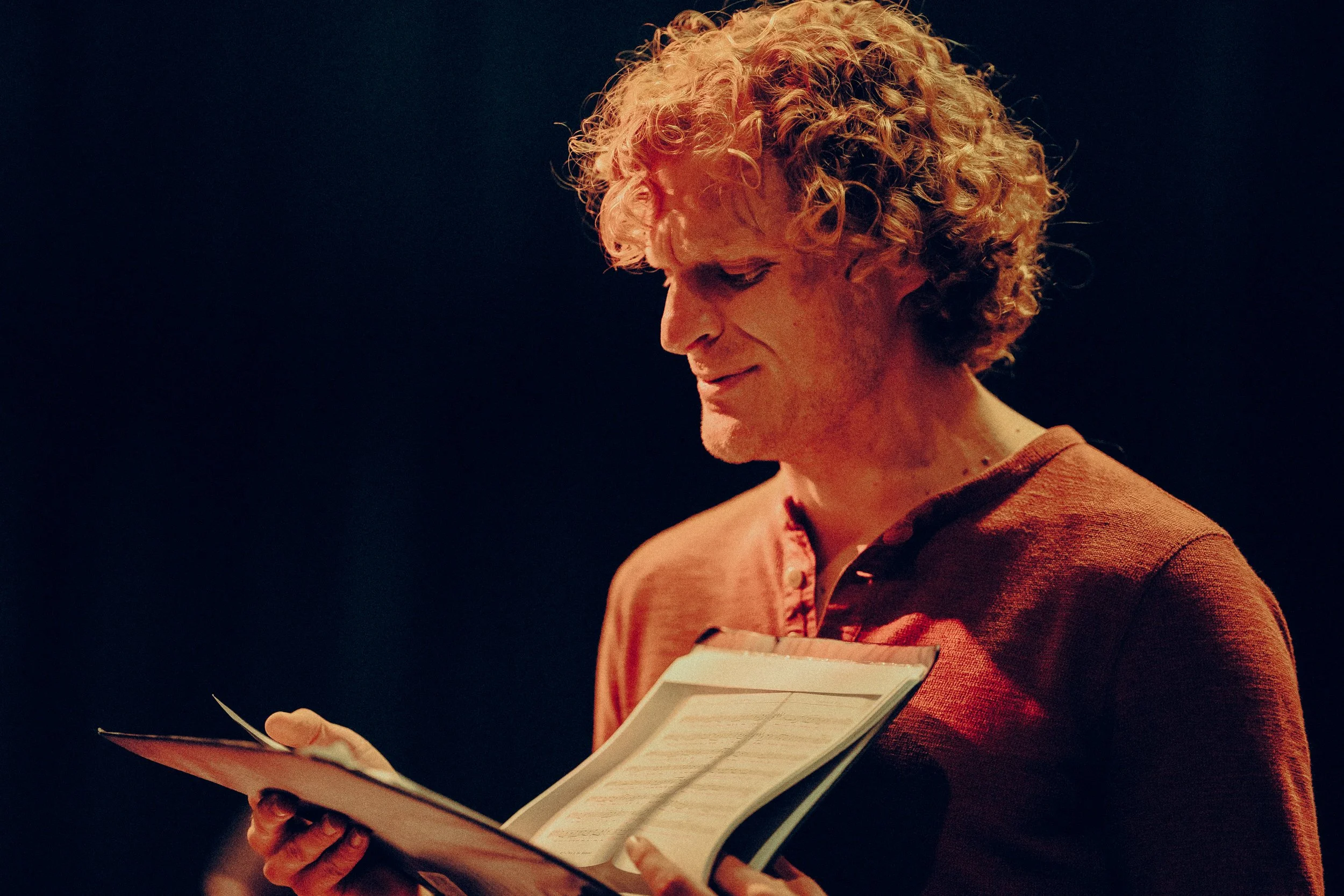 A person looking at sheet music with a thoughtful expression, wearing a red shirt, against a dark background.