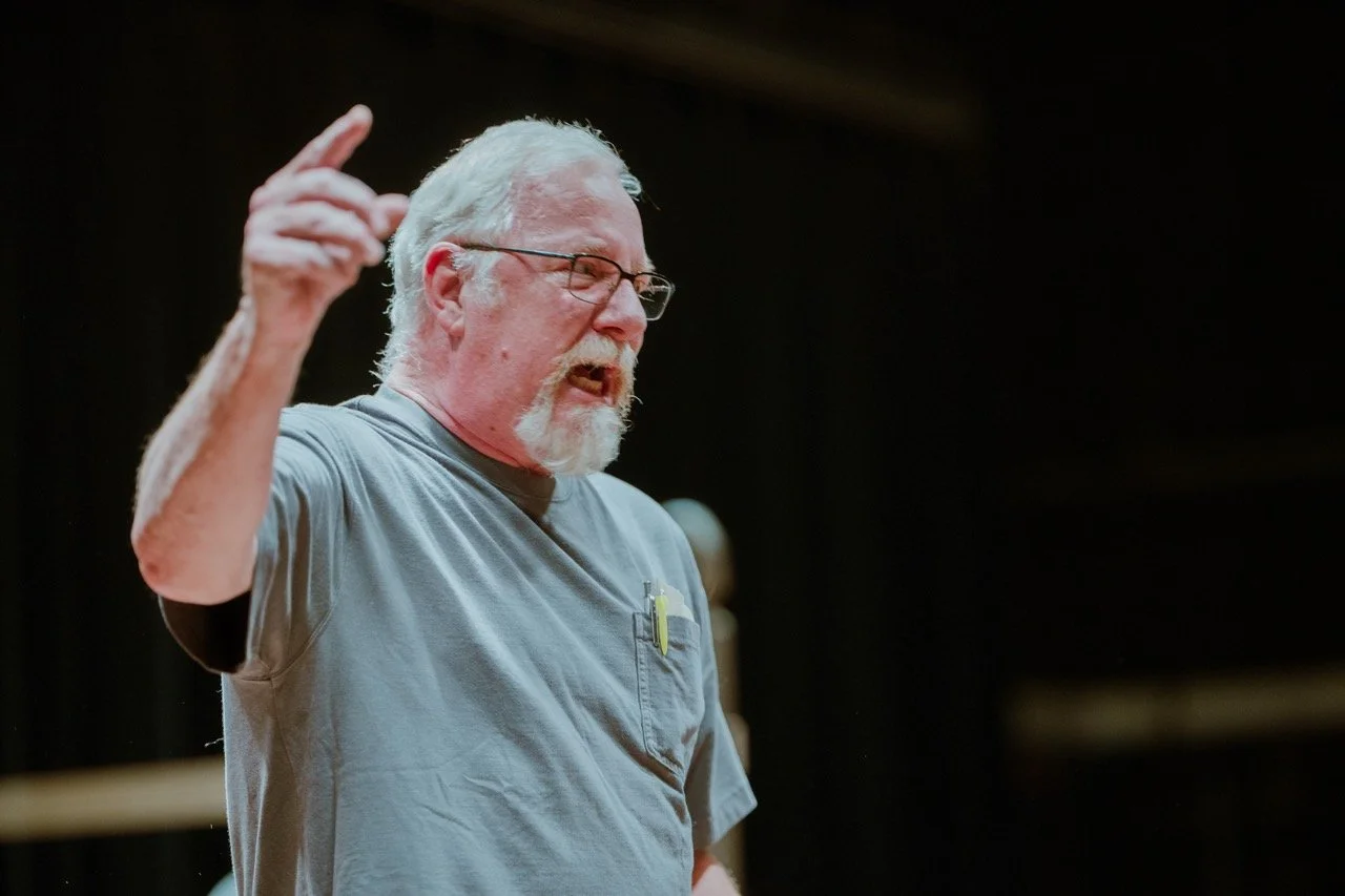An older man with a gray beard, wearing glasses and a gray shirt, is speaking passionately, pointing with one finger.