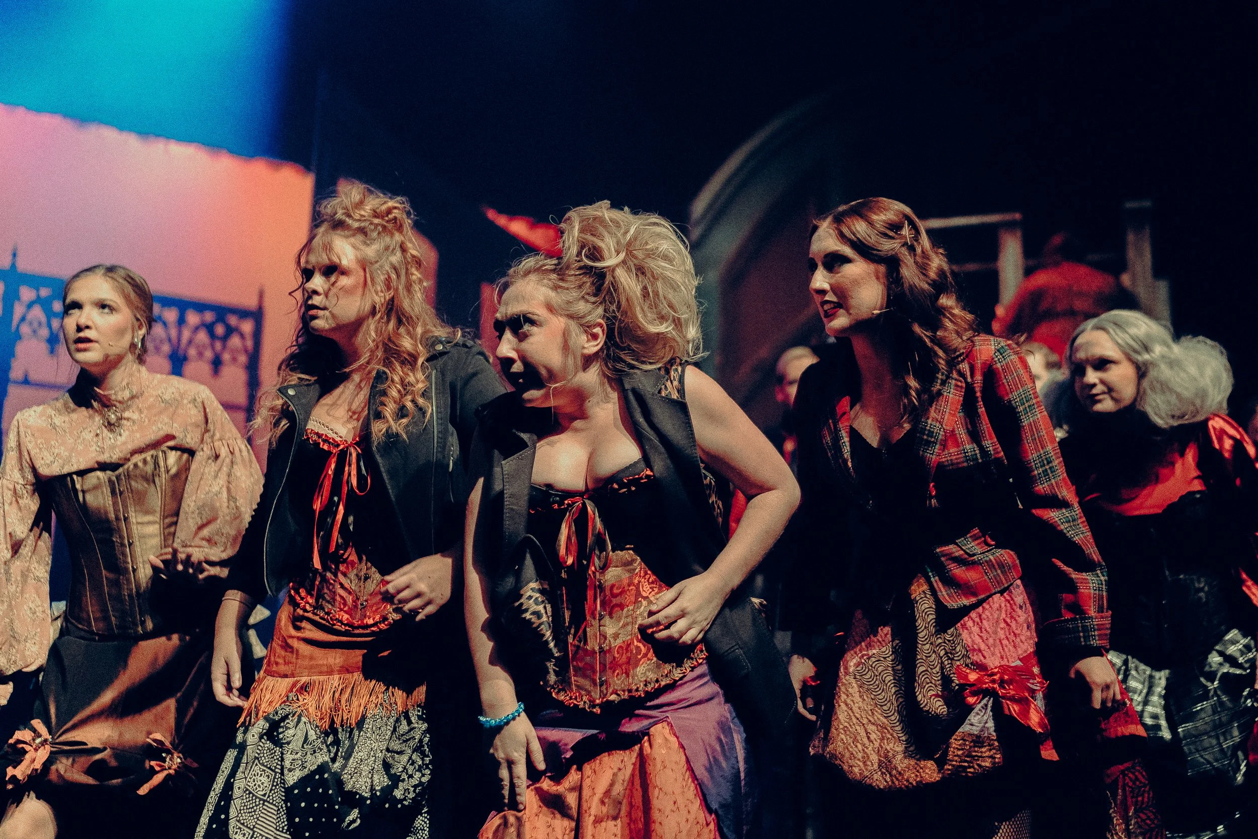 Group of women in theatrical costumes on stage during a performance, with dramatic expressions and lighting.