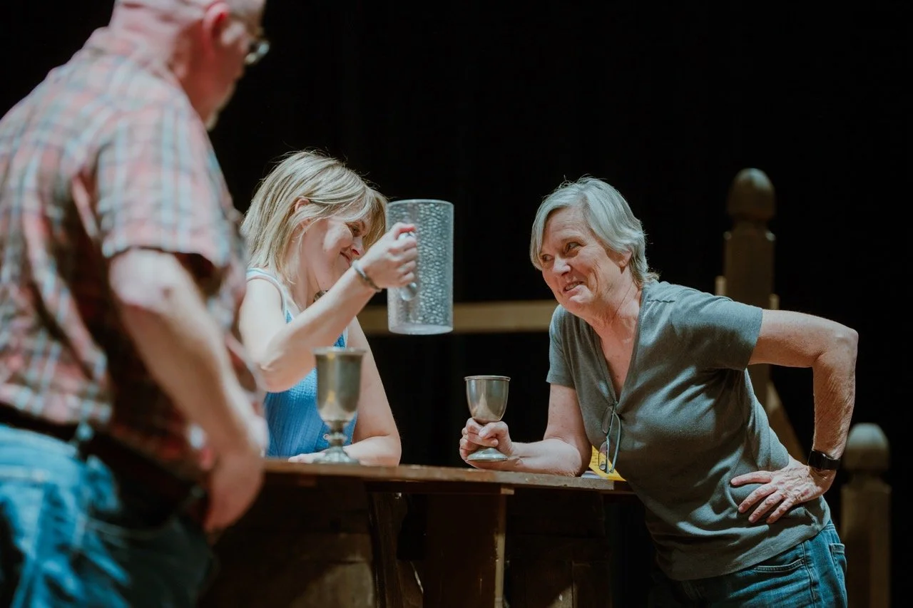 Three people having a friendly conversation at a table, holding metal cups, in a dimly lit setting possibly a bar or pub.