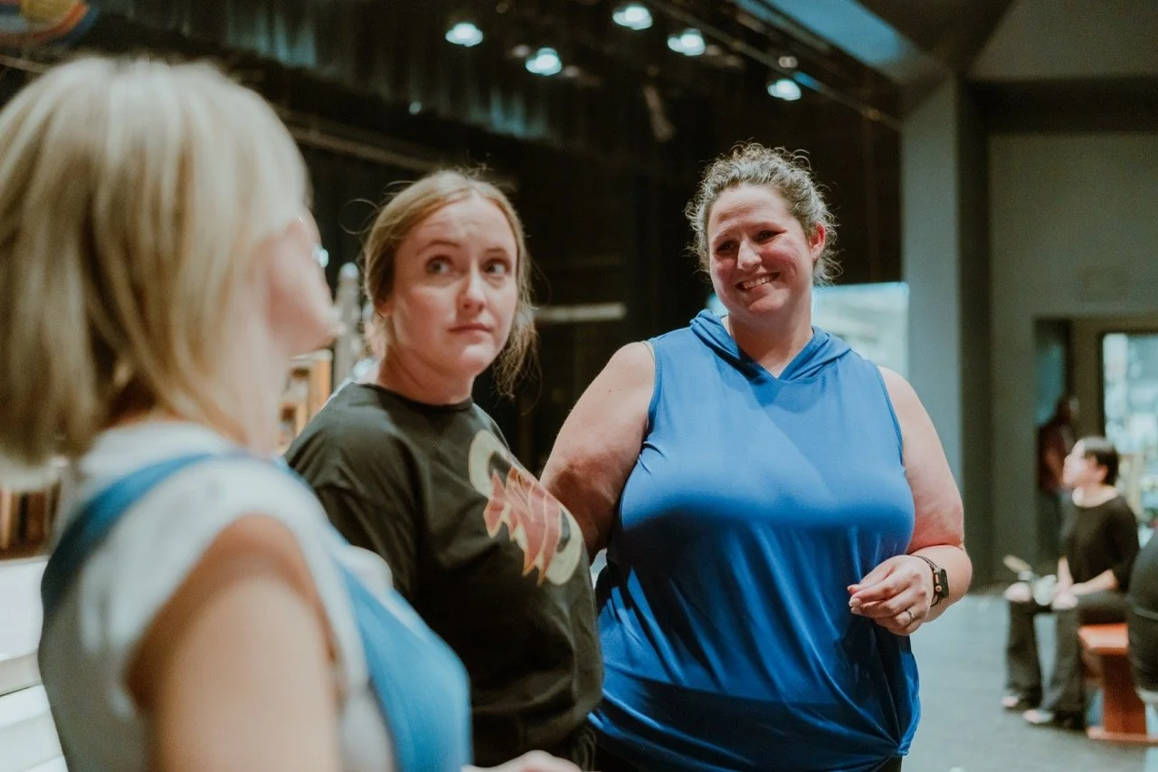 Three women conversing in a dimly lit space, possibly a theater or auditorium. One woman is in focus, smiling and wearing a blue sleeveless top, while the other two are in the foreground and slightly out of focus. Overhead lights are visible.