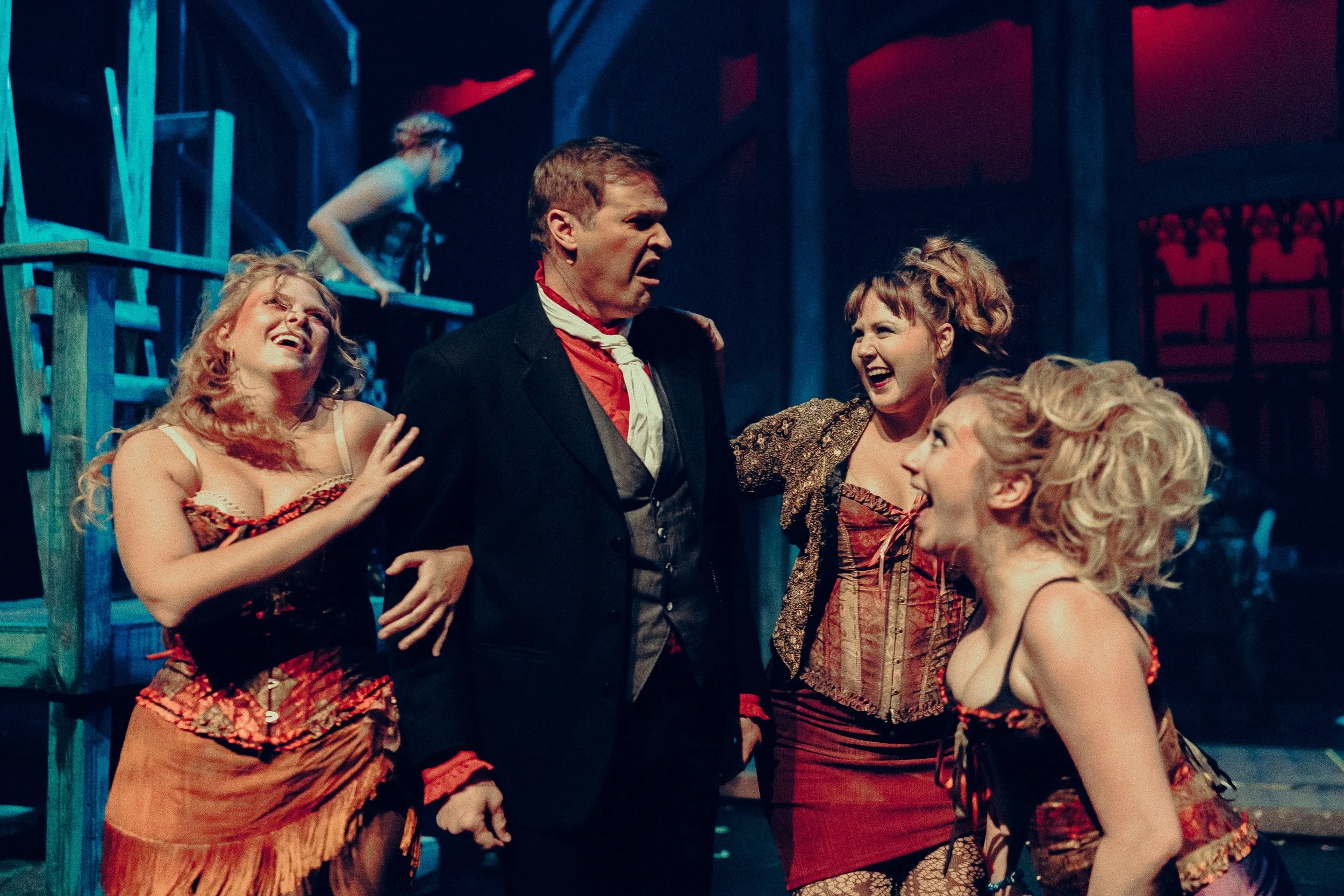 Group of actors in theatrical costumes on stage during a lively performance, featuring Victorian-era attire with corsets and suits, expressing dramatic and cheerful emotions under stage lighting.