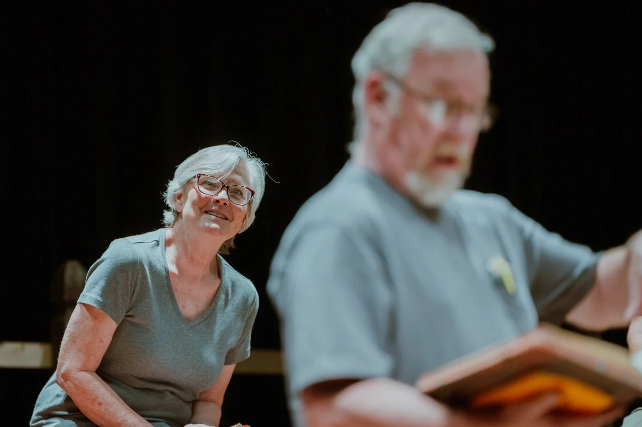 An older woman with short white hair and glasses smiles while sitting, and a blurred man in the foreground appears to be reading a book.