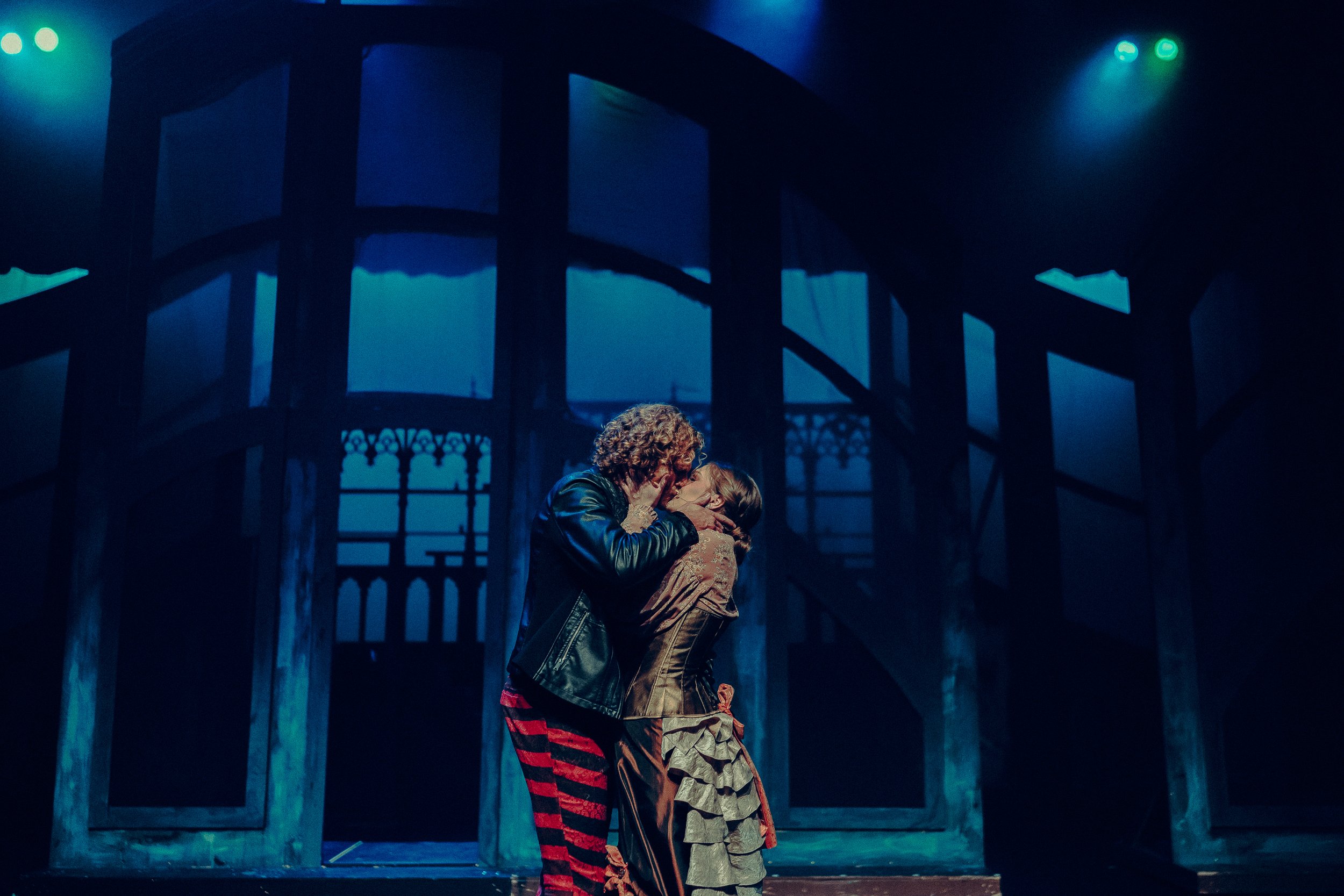Actors performing a dramatic scene on stage with a dimly lit gothic backdrop, wearing theatrical costumes in a dark, moody setting.