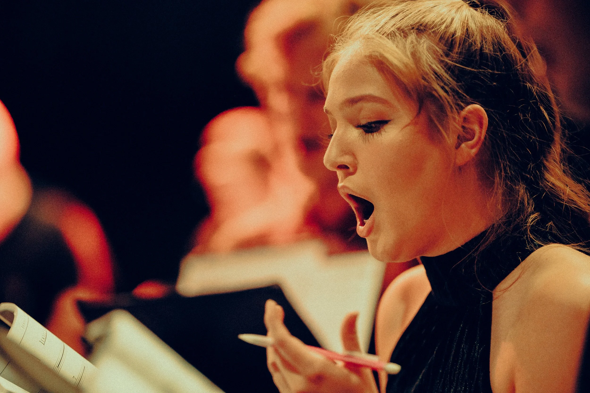 A woman singing passionately, holding a pen and music sheet in a choir or rehearsal setting.