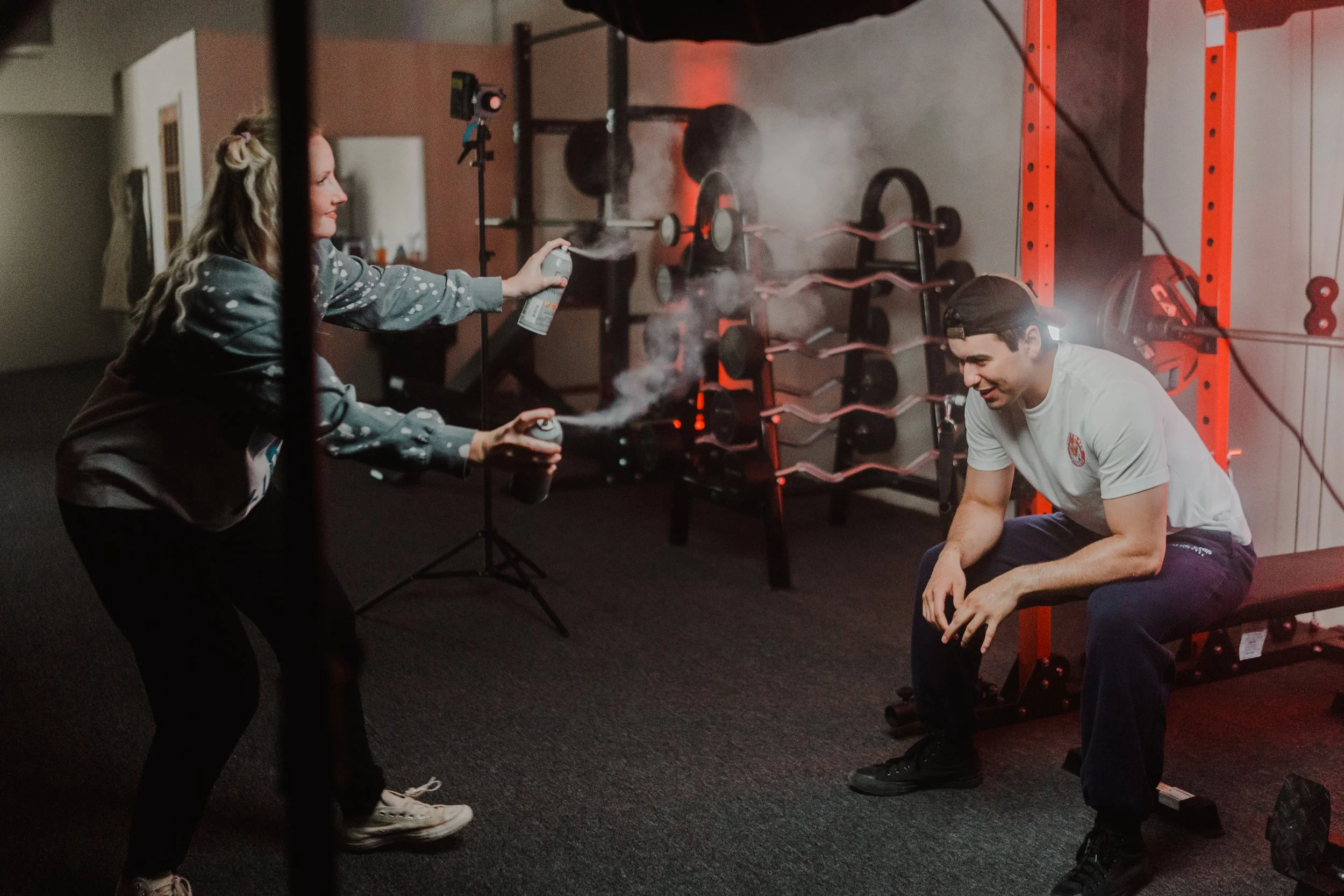 A woman spraying mist towards a seated man in a gym with weights in the background.