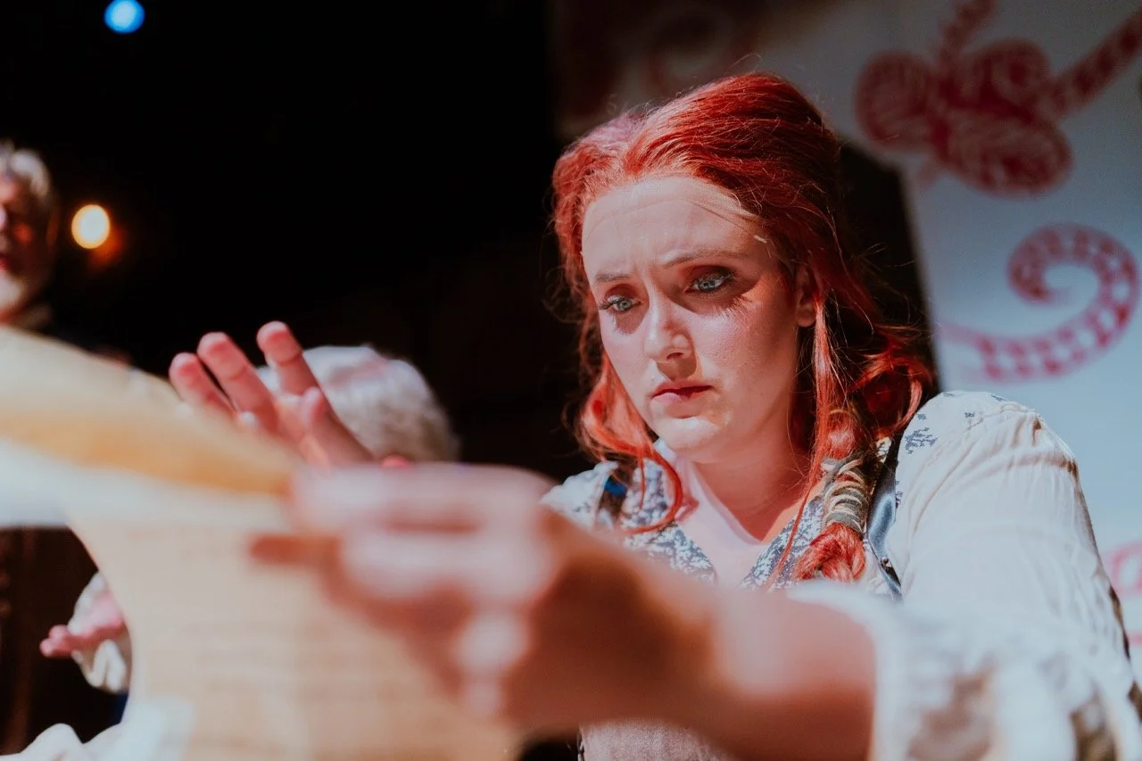 A woman with red hair and braided pigtails looking intently at a document, with a serious expression, in a dimly lit room.