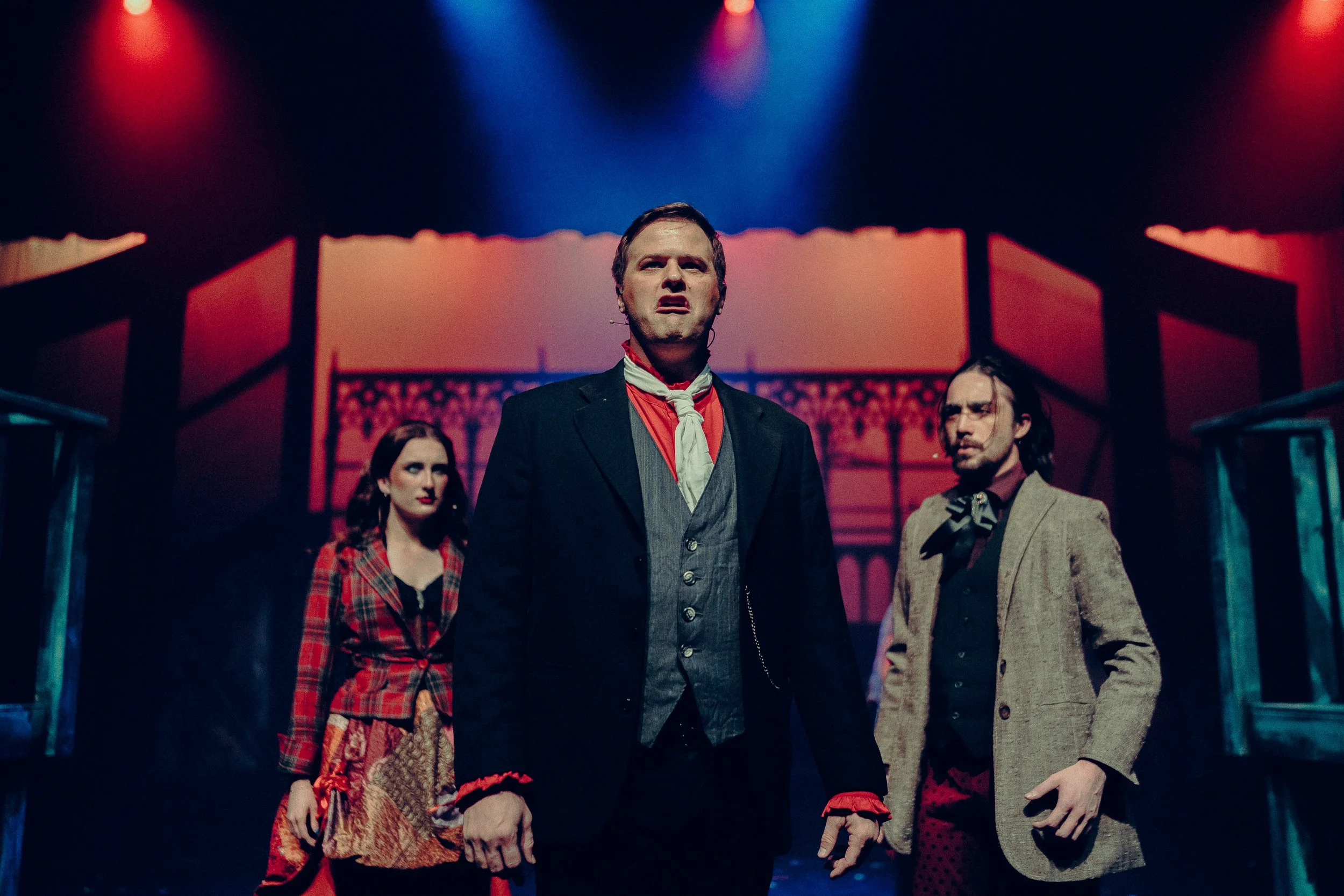 Actors in period costumes performing on a dimly lit theater stage, with red and blue spotlights overhead.