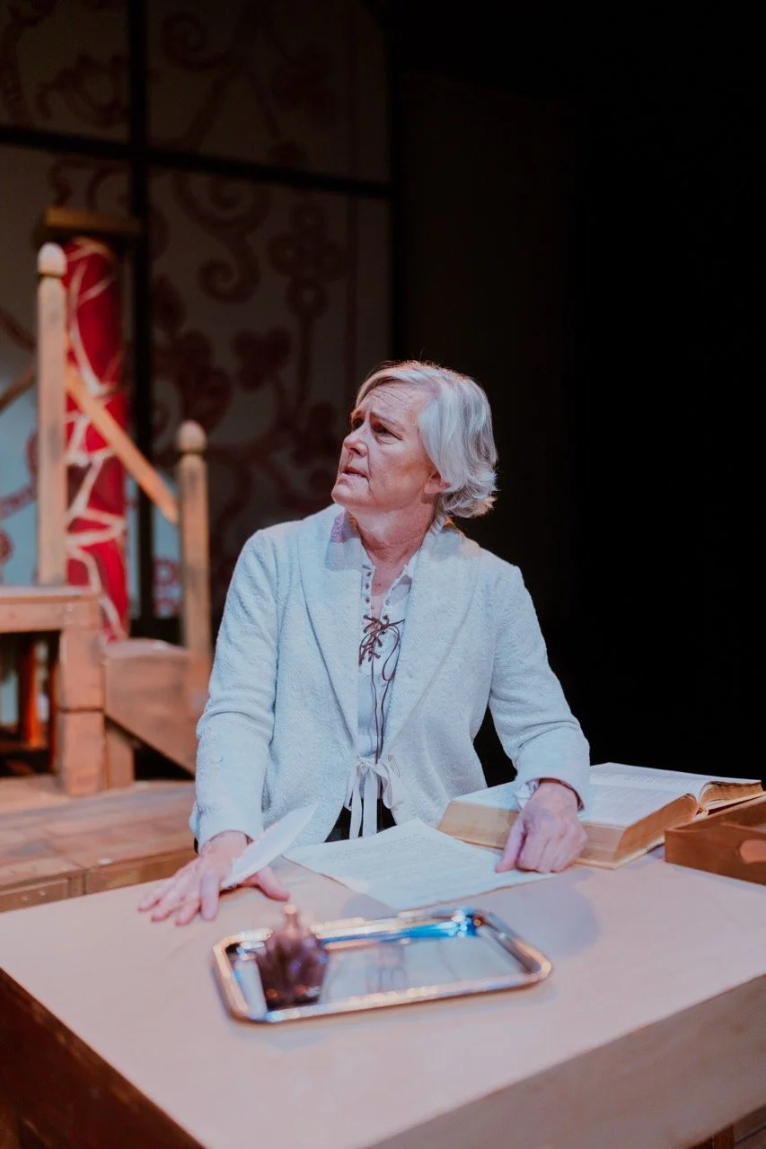 Person in a theater setting, wearing a light jacket, sitting at a wooden desk with open books and a tray with a quill.