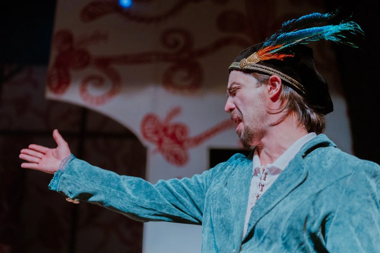 Person wearing a feathered hat and blue costume, gesturing dramatically on stage.