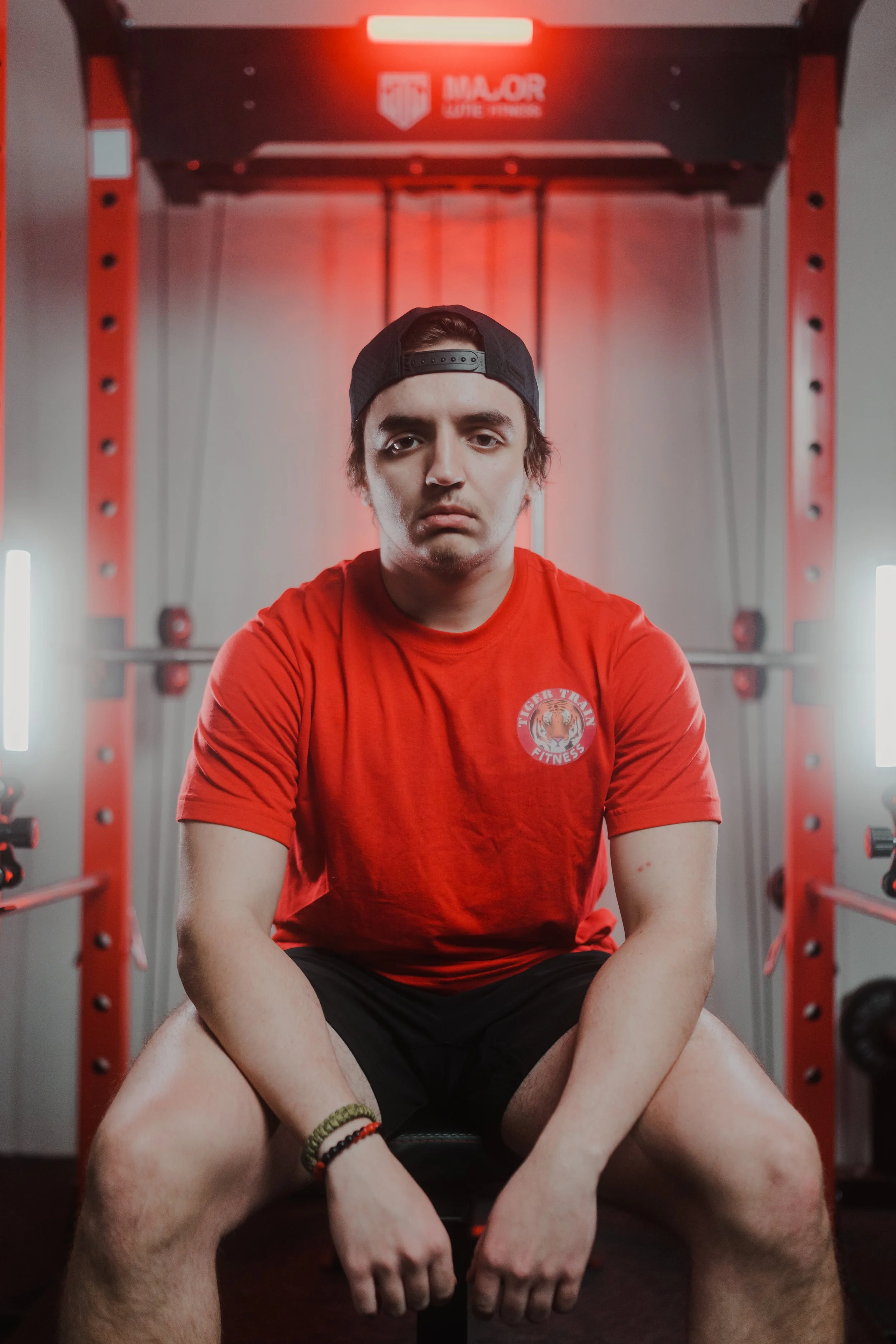 Person in a red "Tiger Tuff Fitness" shirt and cap seated in a gym, surrounded by exercise equipment with red lighting.