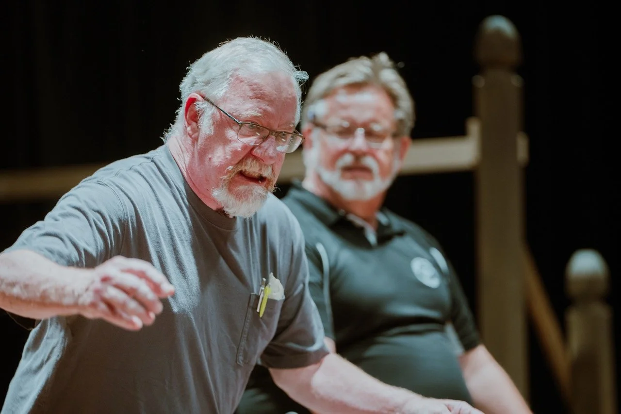 Two older men talking, one gesturing passionately, indoor setting.