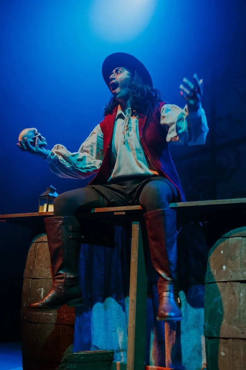 Stage actor in period costume holding a skull, performing under blue lighting.