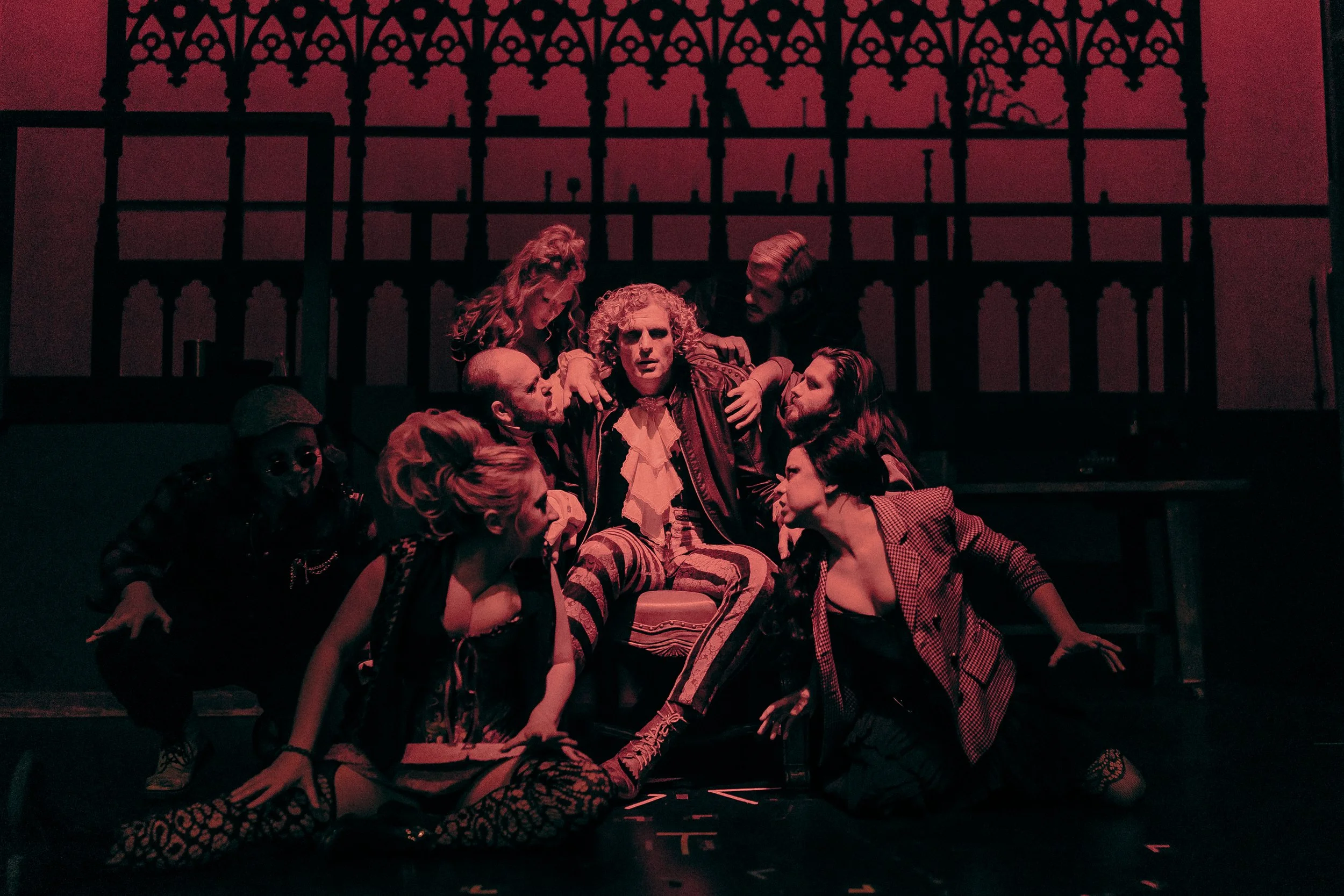 Group of performers in theatrical scene with Gothic backdrop, atmospheric red lighting, and diverse costumes.