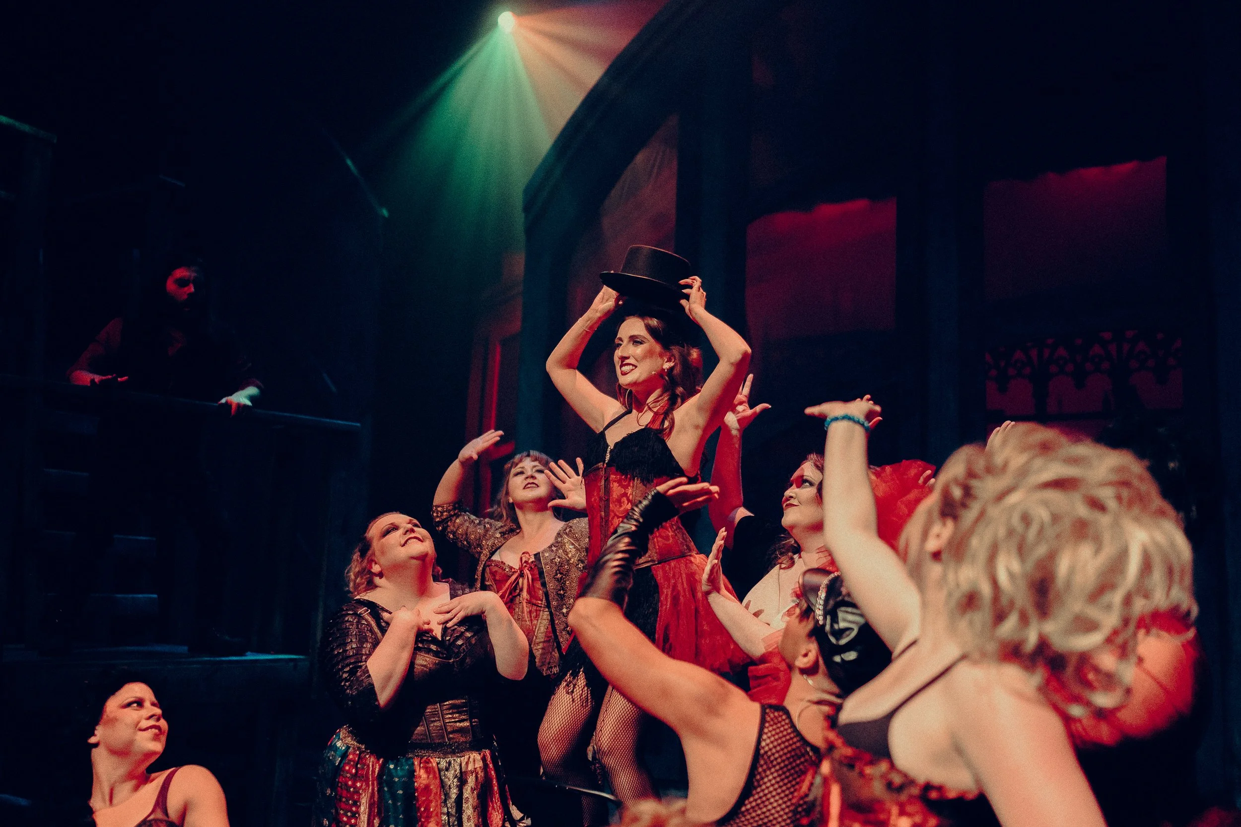 Performers in a theatrical scene, one holding a top hat and wearing a corset-style costume, surrounded by others in similar attire, with stage lighting and a dark, atmospheric background.