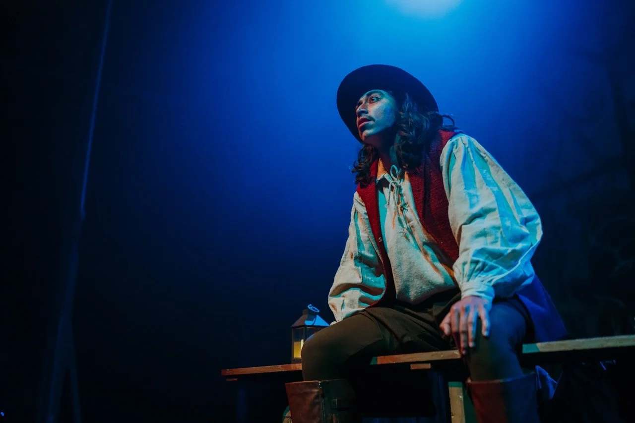 Person in period costume with hat sitting on a wooden bench under blue stage lighting.