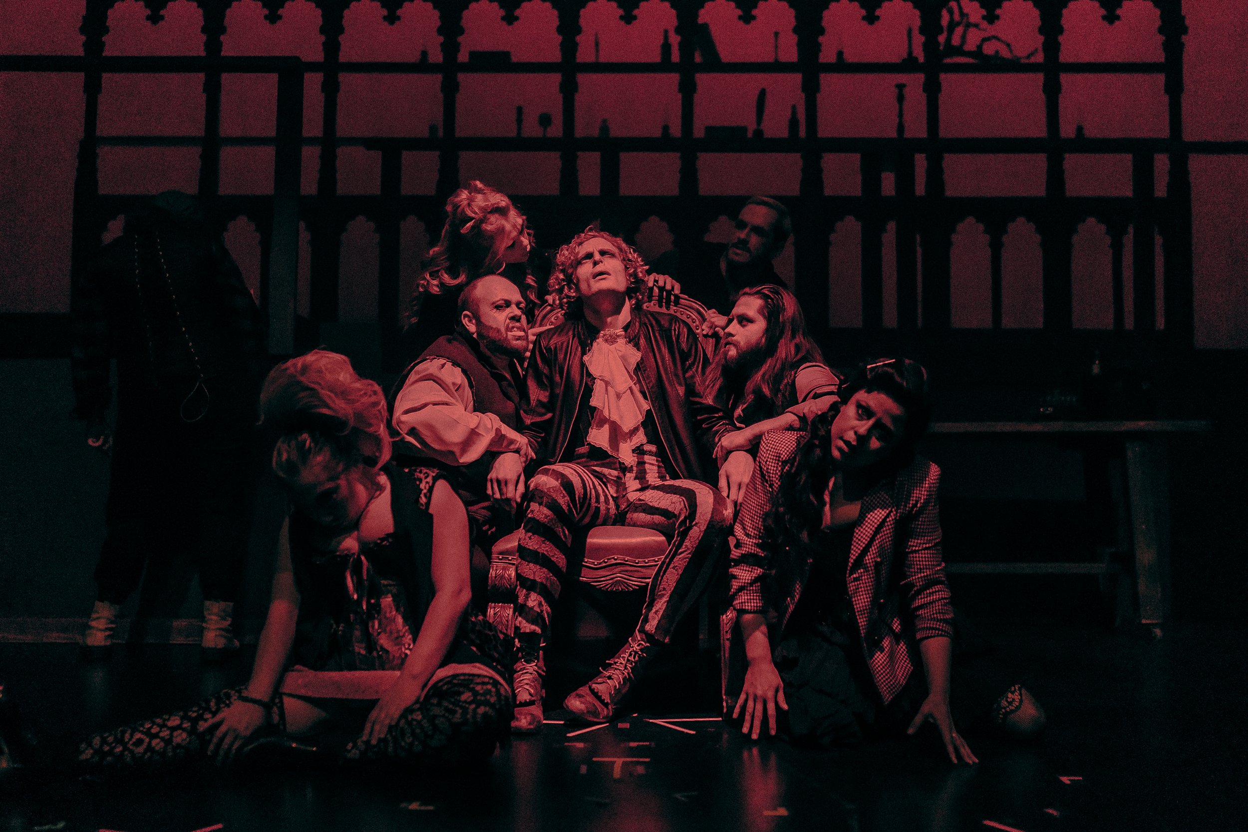 A group of performers on stage in a theatrical scene with dramatic red lighting. One person is seated on a chair wearing striped pants, surrounded by others in various poses. The background features a silhouette resembling gothic architecture.