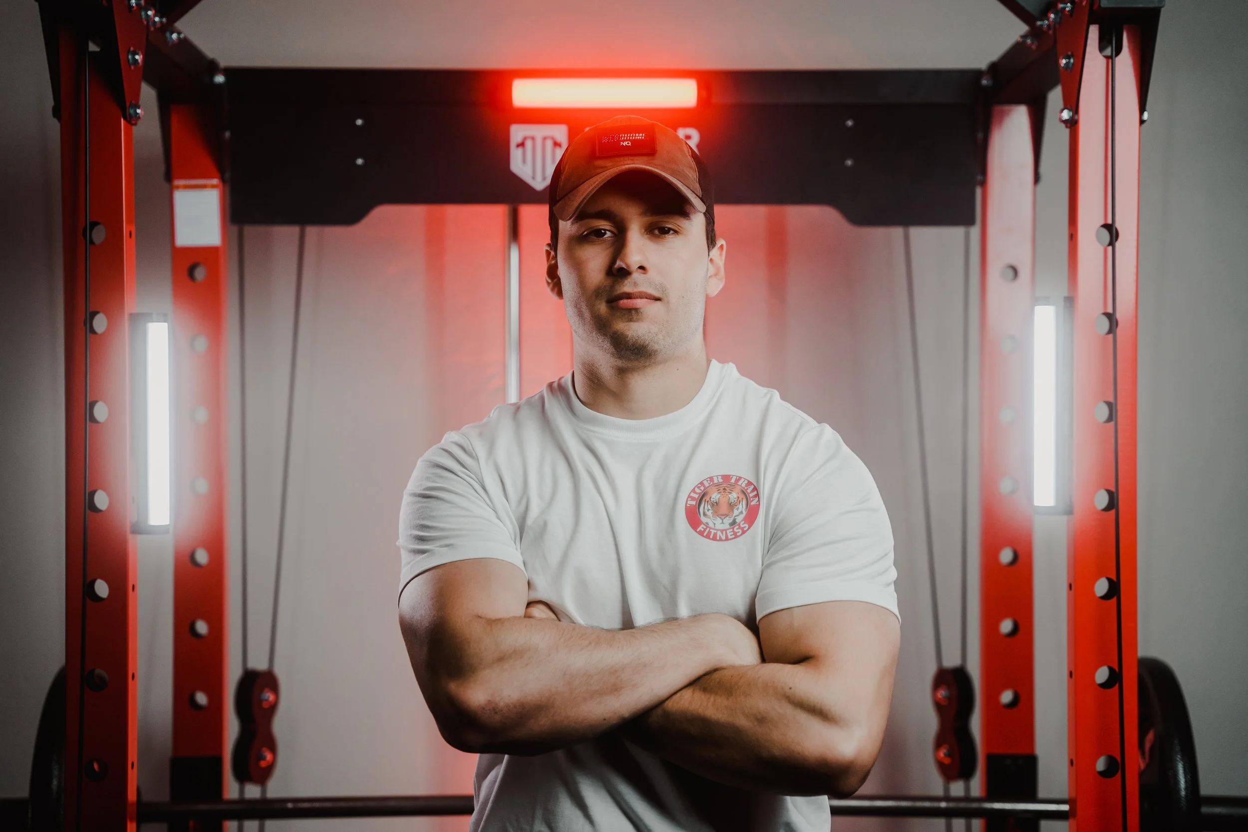 Man in white t-shirt and cap standing in a gym with red equipment and arms crossed.