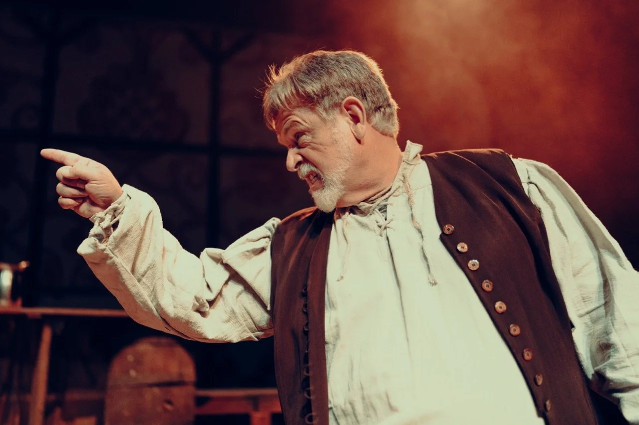 An older man in period costume performing on stage, looking angry and pointing his finger.