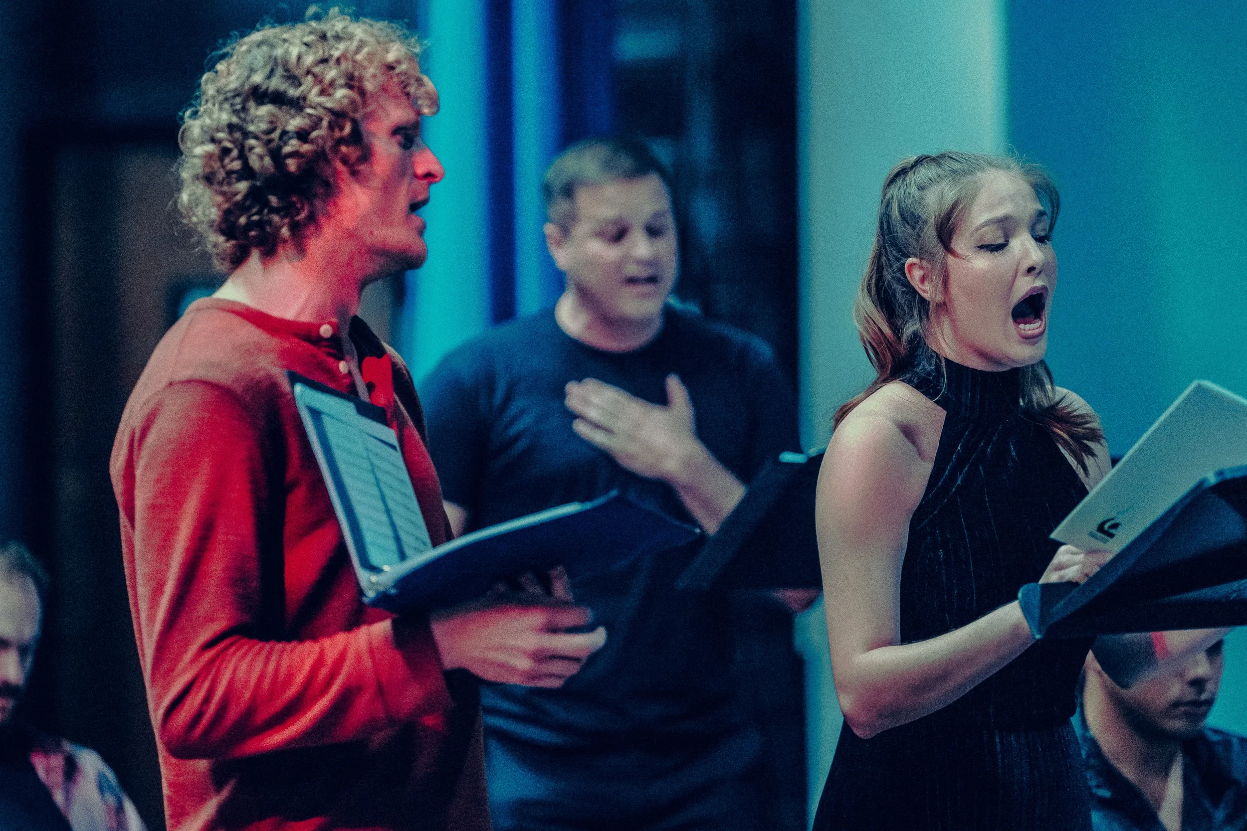 A group of people singing in rehearsal, holding music folders, with one man wearing a red shirt and a woman in a dark dress.