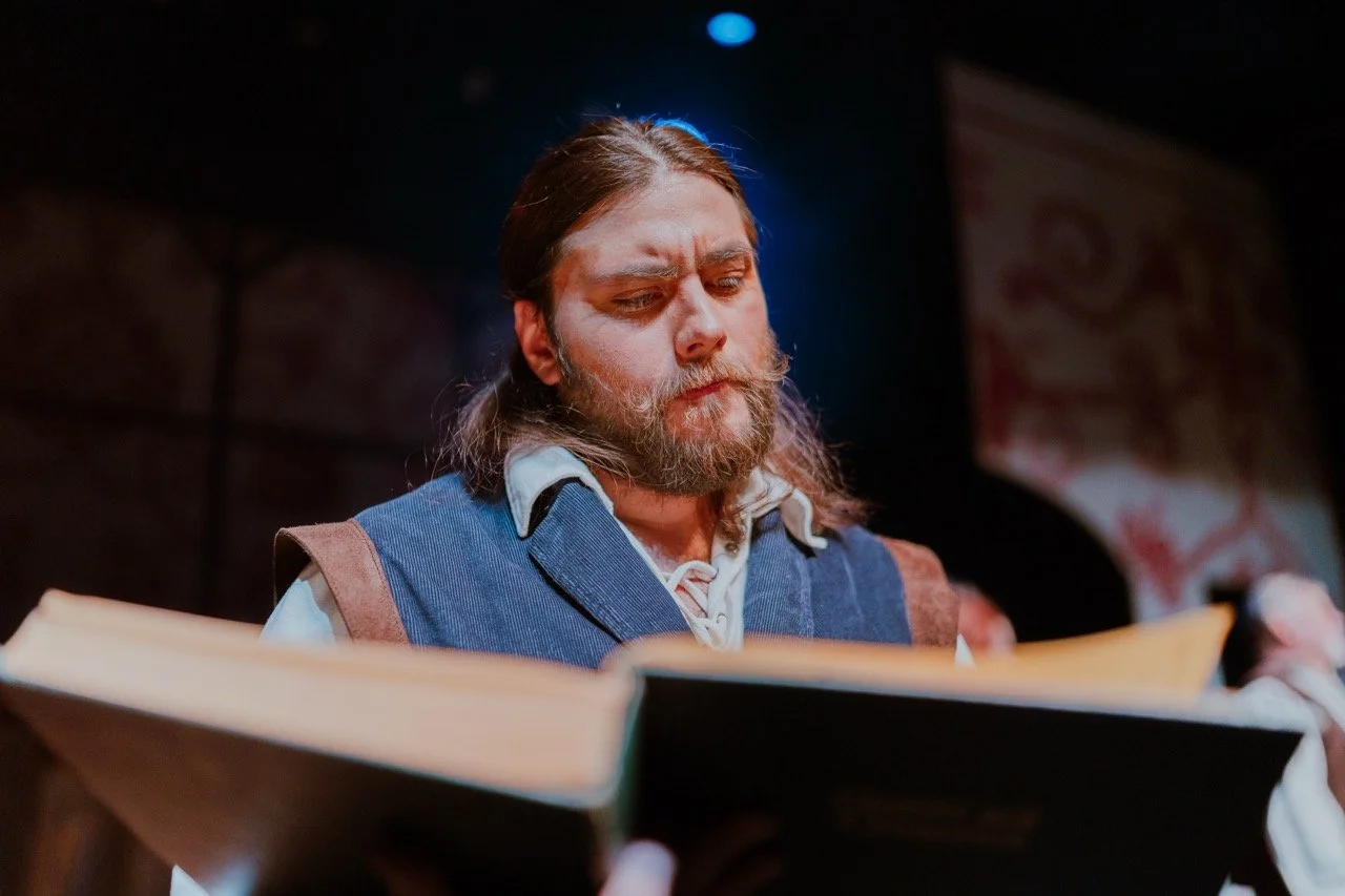 Bearded man in historical costume reading a large book under stage lights.