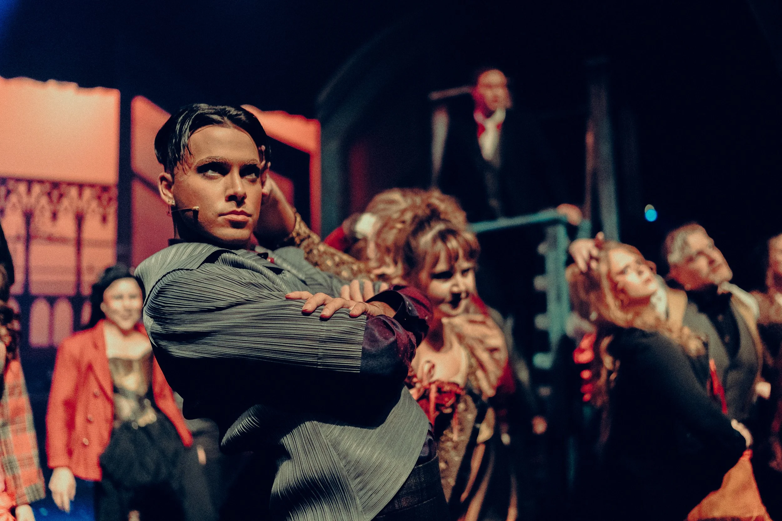 A dramatic stage performance with an actor in the foreground striking a confident pose, surrounded by other performers in vibrant costumes.