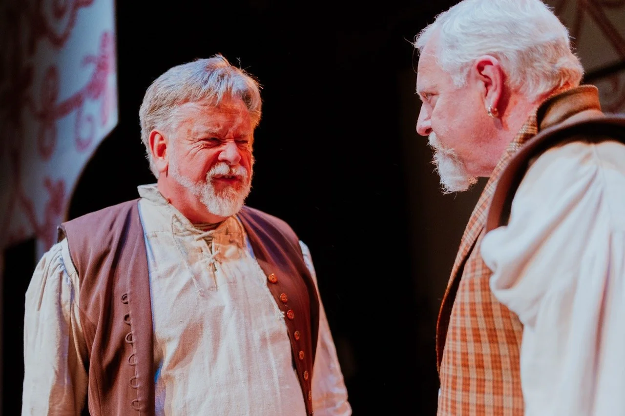 Two older men in period costumes performing on stage, with one man looking serious and the other making a dramatic expression.