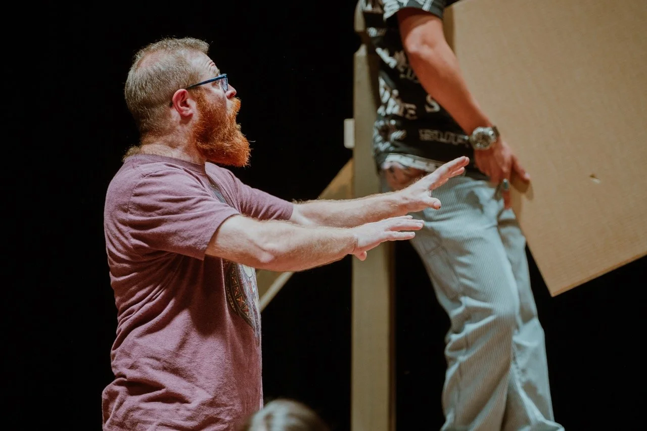 Man with a beard gesturing expressively during a performance or rehearsal, another person holding a cardboard sheet in the background.