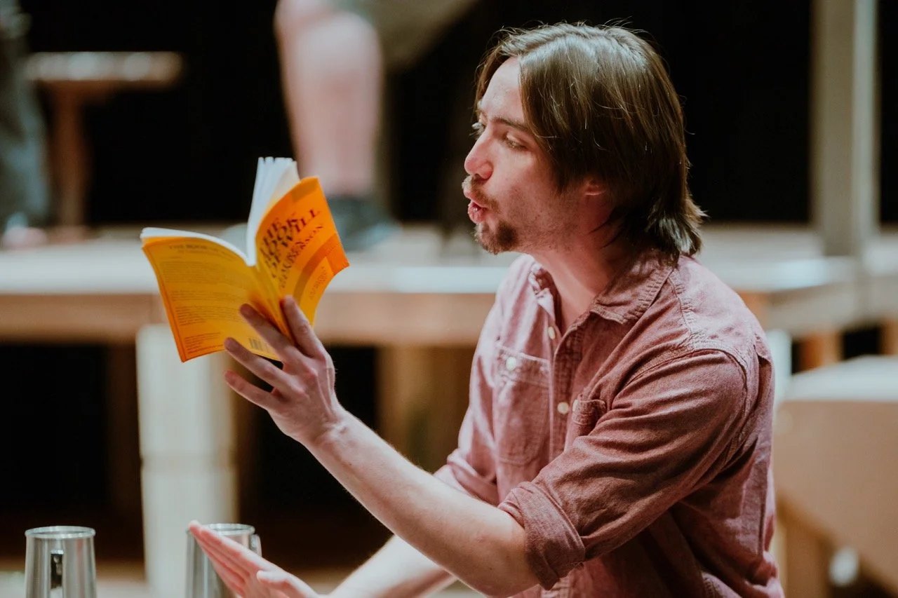 Person reading aloud from a book with an orange cover while sitting inside a room.