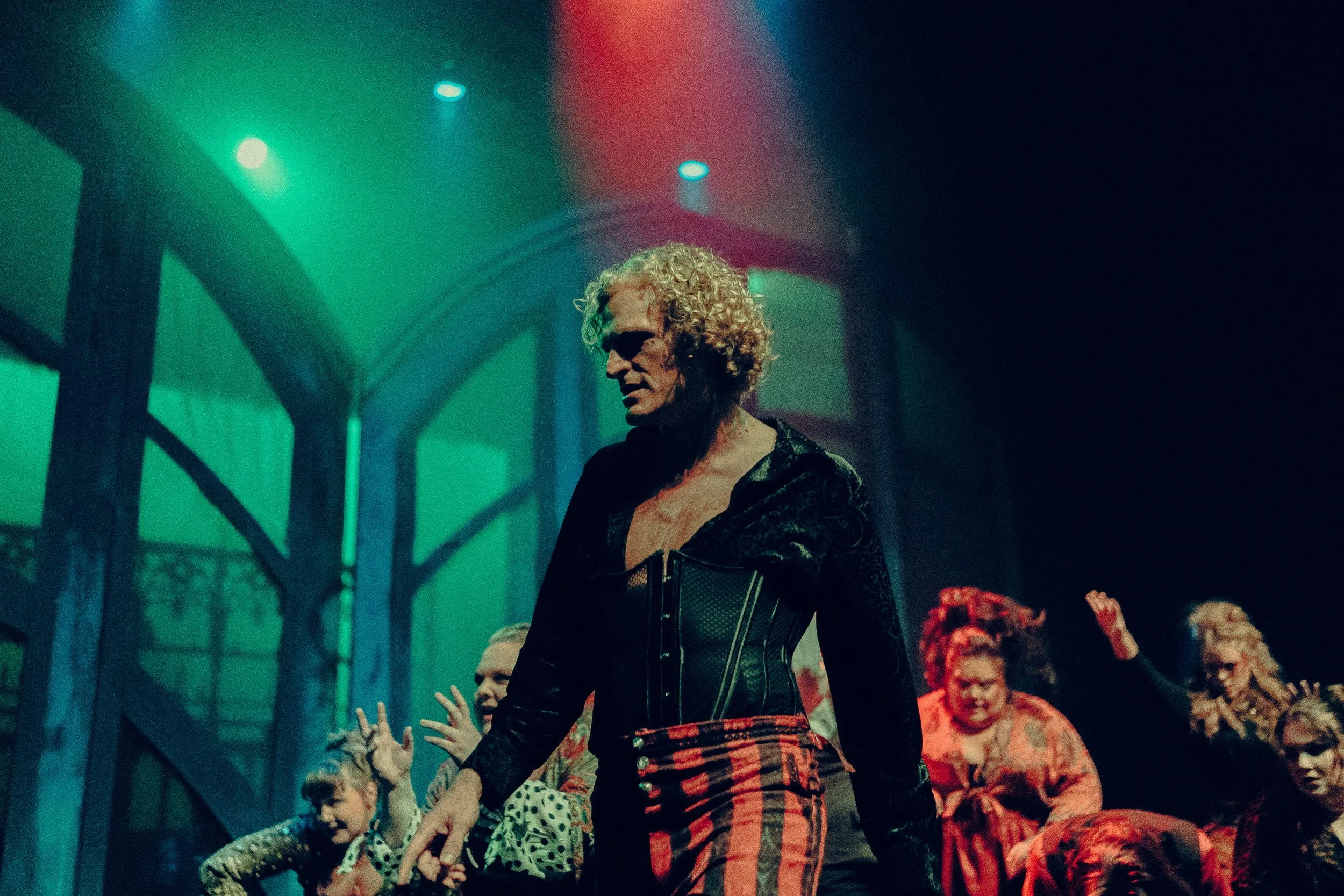 A theatrical stage performance with a person in a black velvet top and red striped pants, surrounded by other performers in dramatic poses under colored lighting.