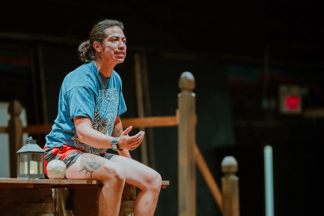 Person sitting on stage, wearing a blue shirt, shorts, and a wristwatch, gesturing expressively. A lantern and a sphere are placed beside them. Wooden railings and a dark background are visible.