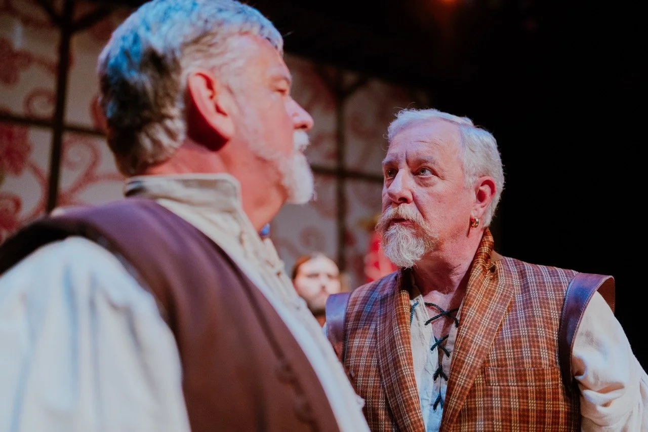 Two older men in historical costumes, one facing the camera and looking serious, the other seen from the side, involved in a theatrical scene inside a decorated venue.