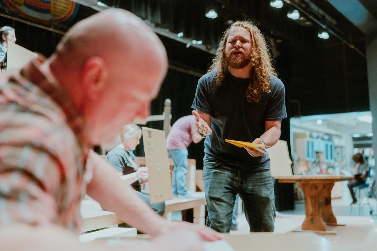 People in a theater setting, with one person holding a script, possibly rehearsing a scene. Background includes stage equipment and other individuals in the area.