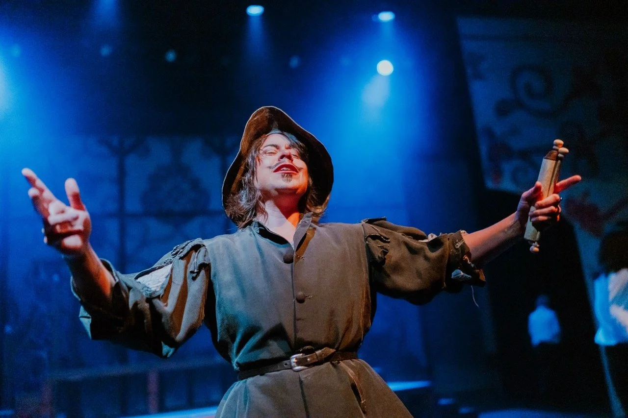 A person in a theatrical costume with a wide-brimmed hat and dark robe, performing on stage with an expressive gesture. They hold a scroll in one hand and are lit by dramatic blue stage lighting.