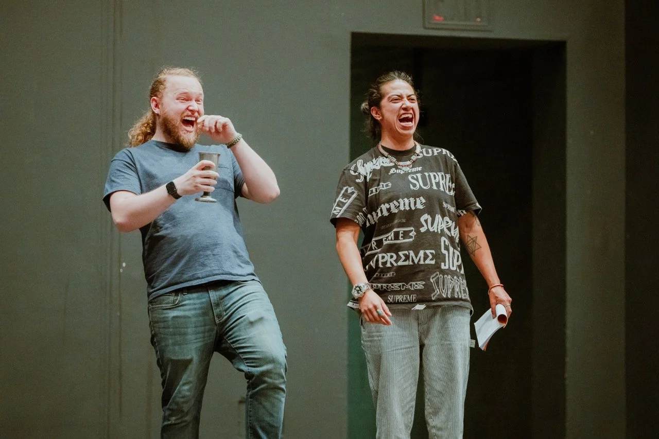 Two people standing on stage, one holding a goblet and the other with a paper. Both are laughing and wearing casual clothes.