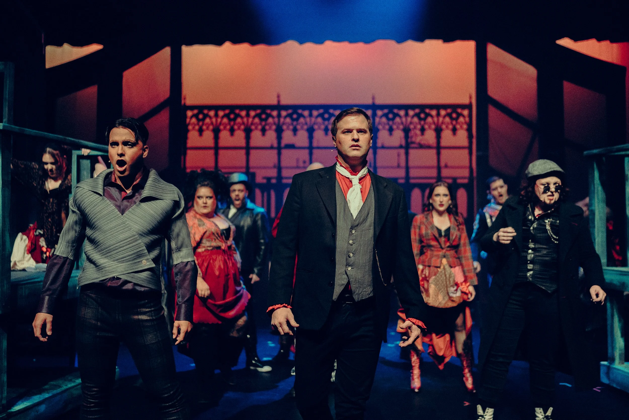 Theatrical performance with diverse cast in dramatic costumes on stage, dark lighting, ornate backdrop, intense expressions.