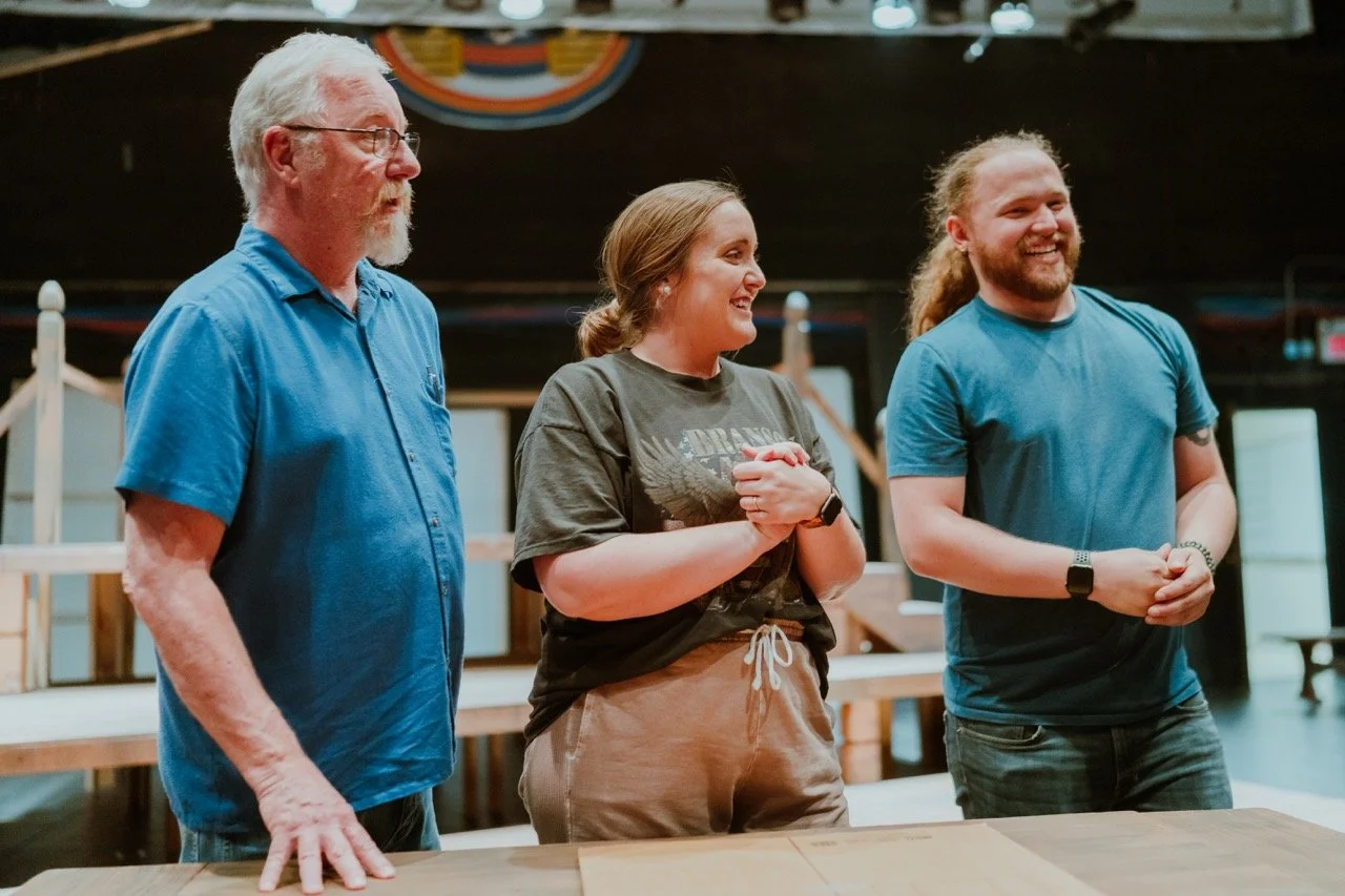 Three people standing indoors, smiling and looking focused, with tables and staging in the background.