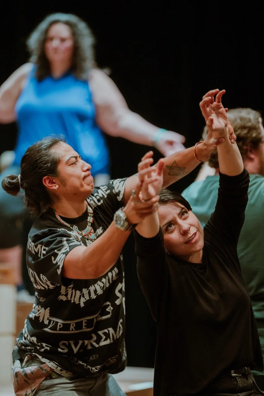 A group of people performing expressive movements on stage. Two individuals in the foreground are intensely focused, with one aiding the other's arm stretch. They wear casual clothing with bold patterns and patterns on black fabric.