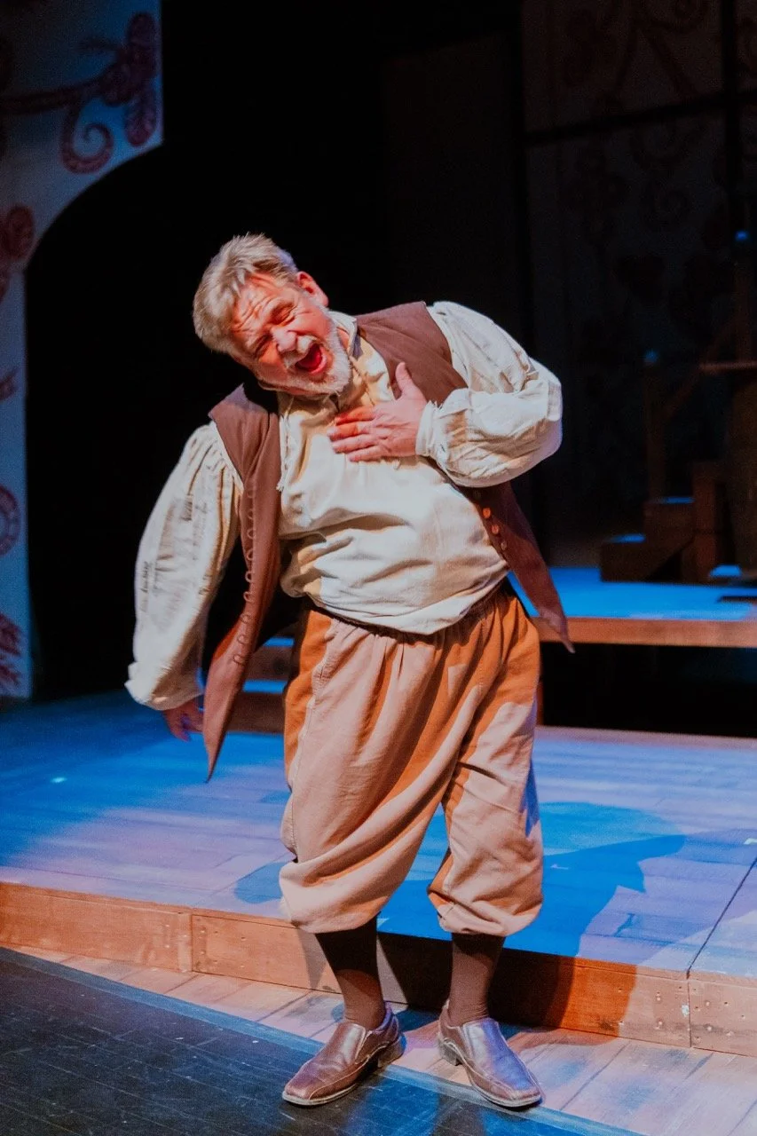 Actor performing on stage in medieval costume with exaggerated expression, wooden floor, theater setting.